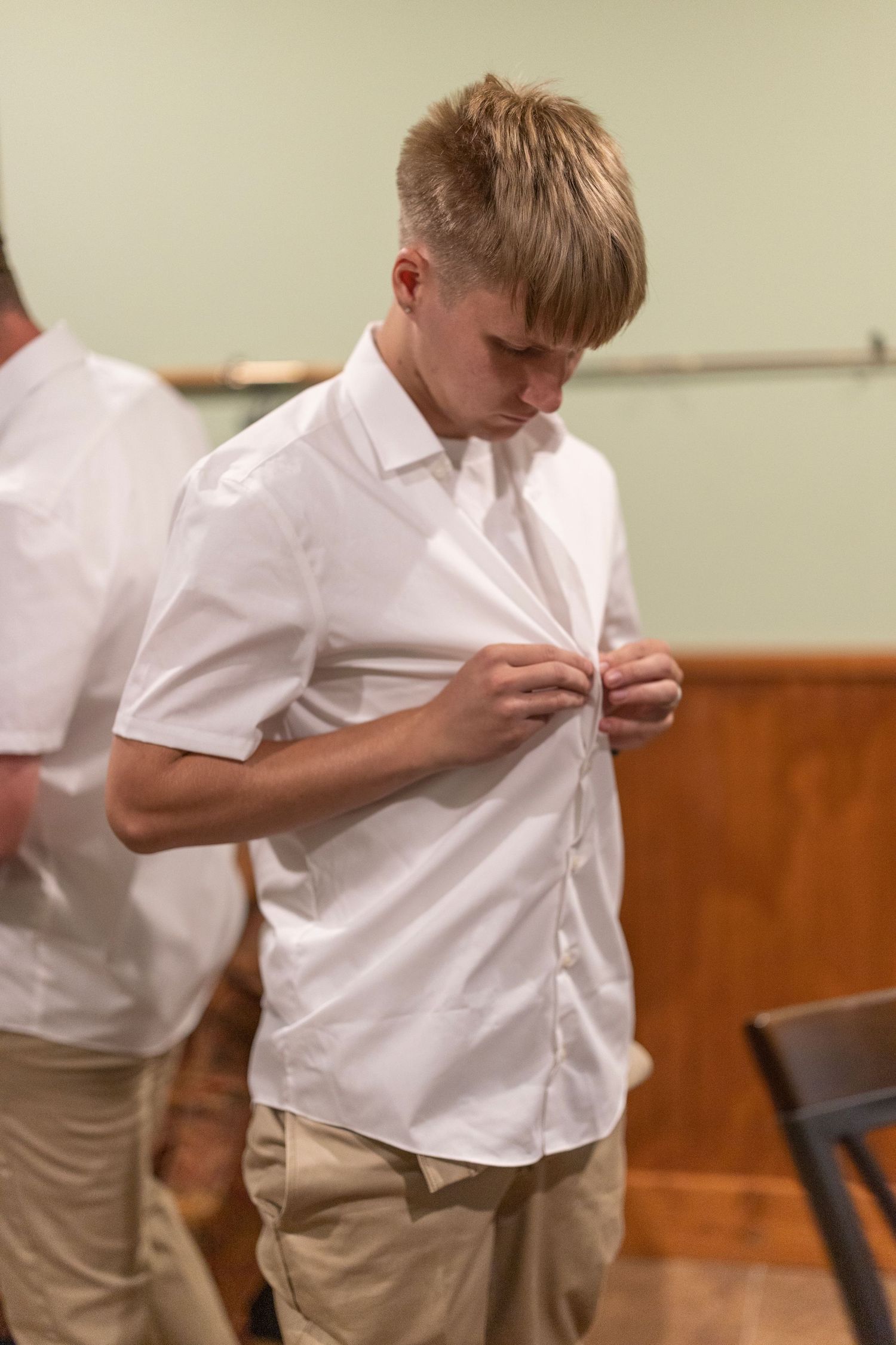 Young teen boy buttoning white short-sleeve dress shirt while getting ready, wearing khaki pants indoors.