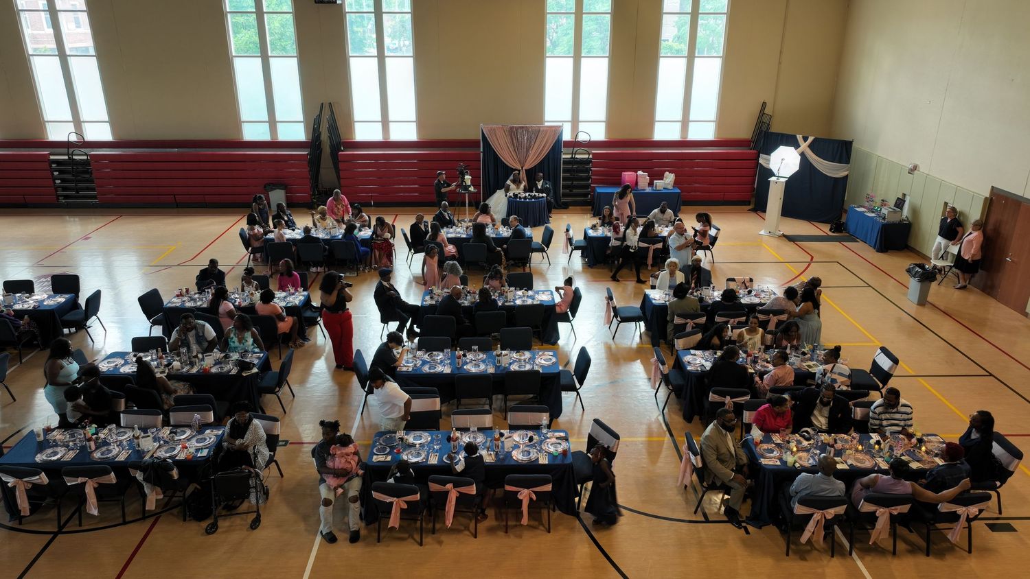 Aerial view of a school gymnasium set up for a banquet with round tables, blue centerpieces, and seated guests.