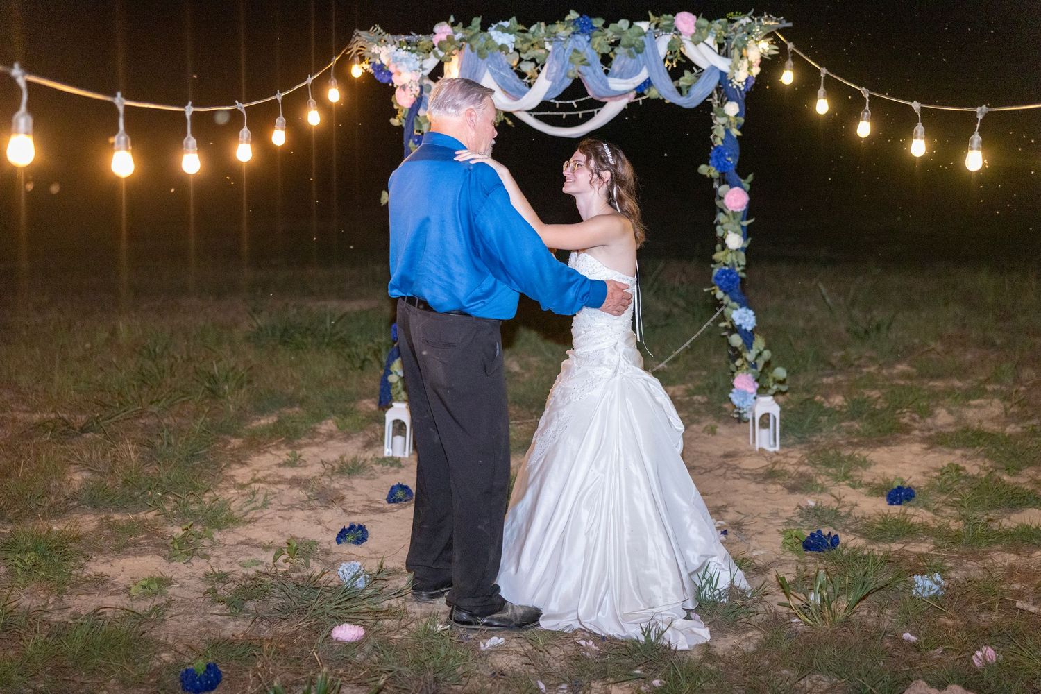 Bride and groom share first dance under floral arch with string lights at outdoor evening wedding ceremony.
