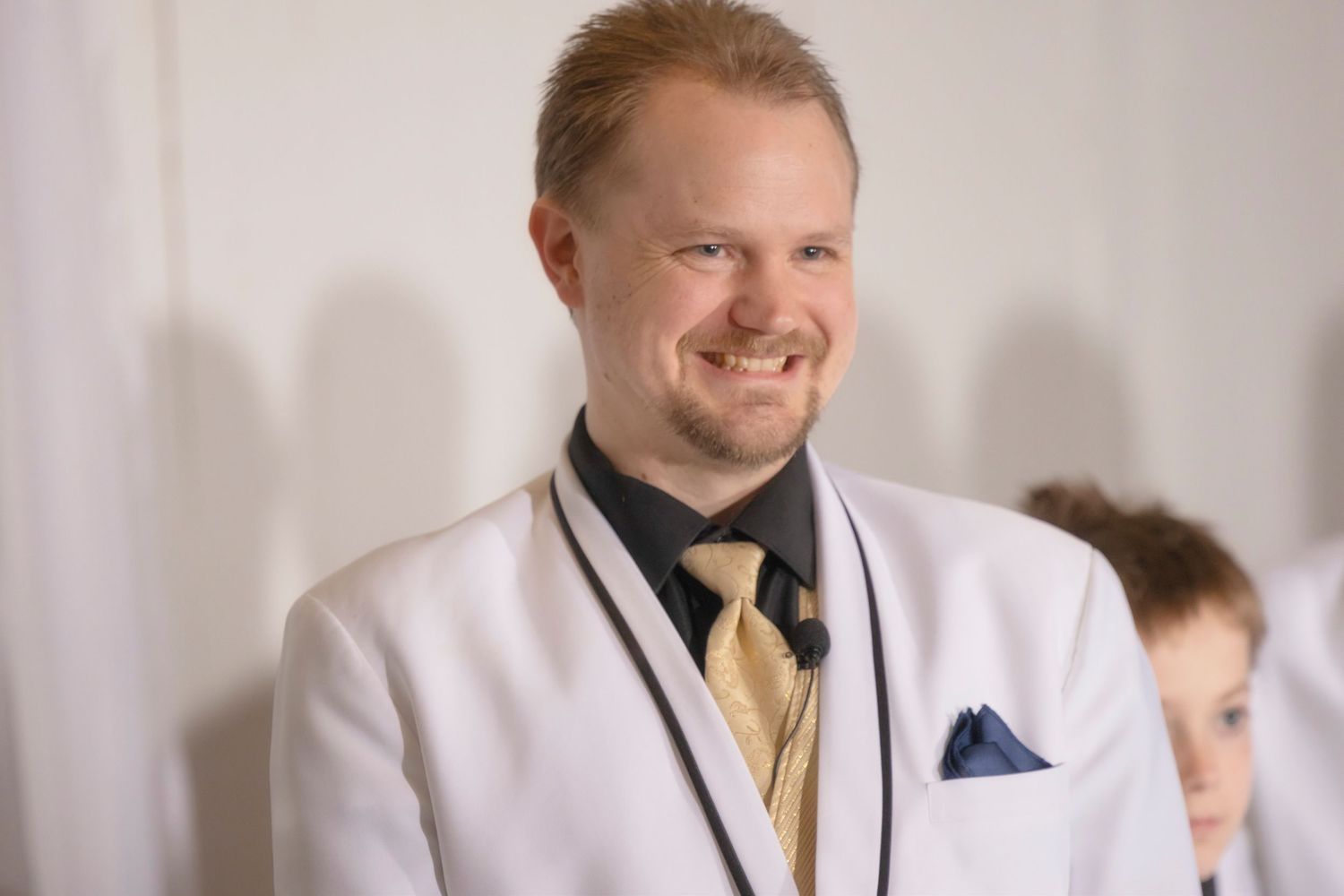 Smiling man in white blazer, black shirt, and gold tie at a formal event.