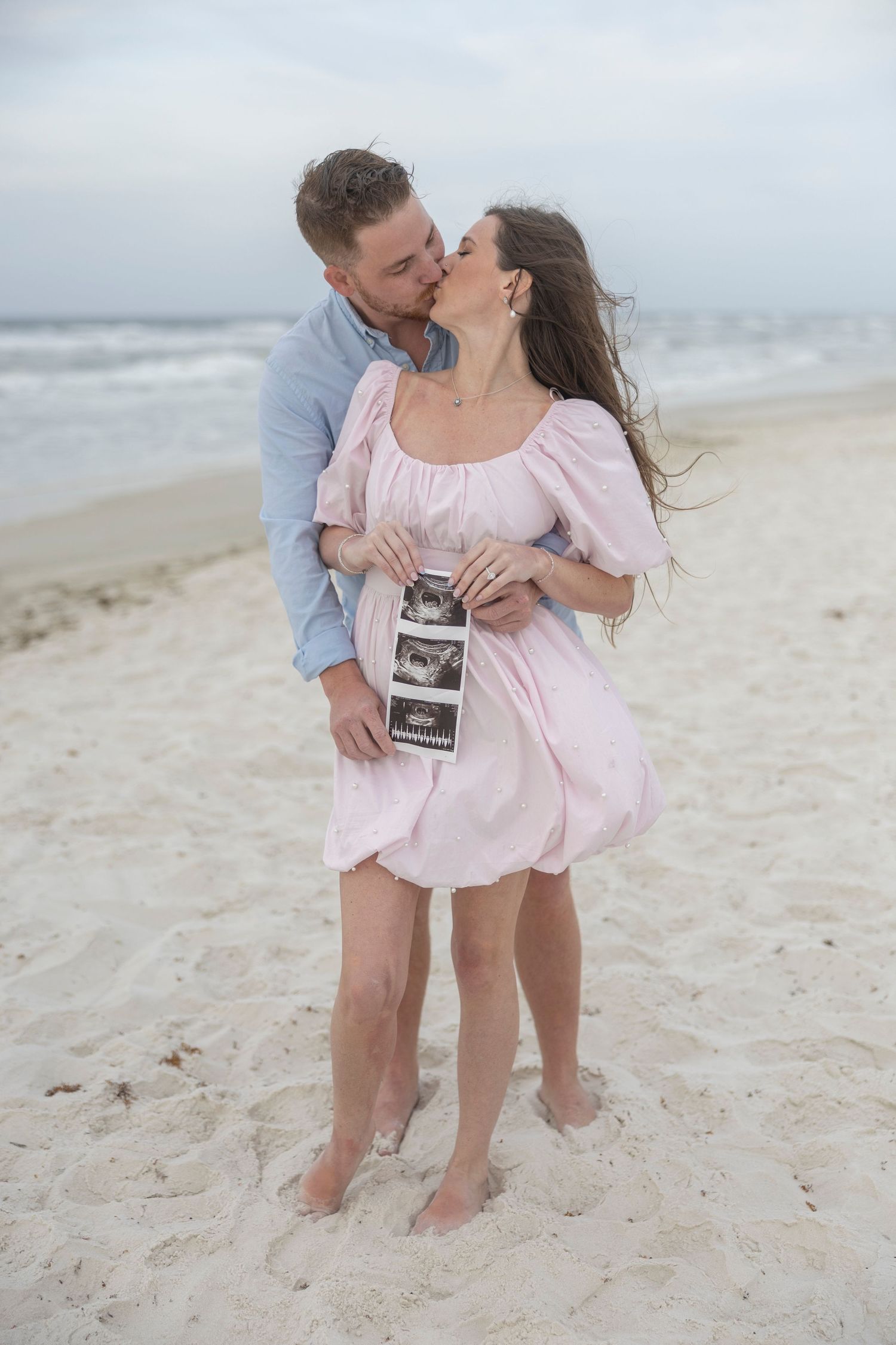 Couple kissing on beach holding ultrasound photos, woman in pink dress announcing pregnancy.