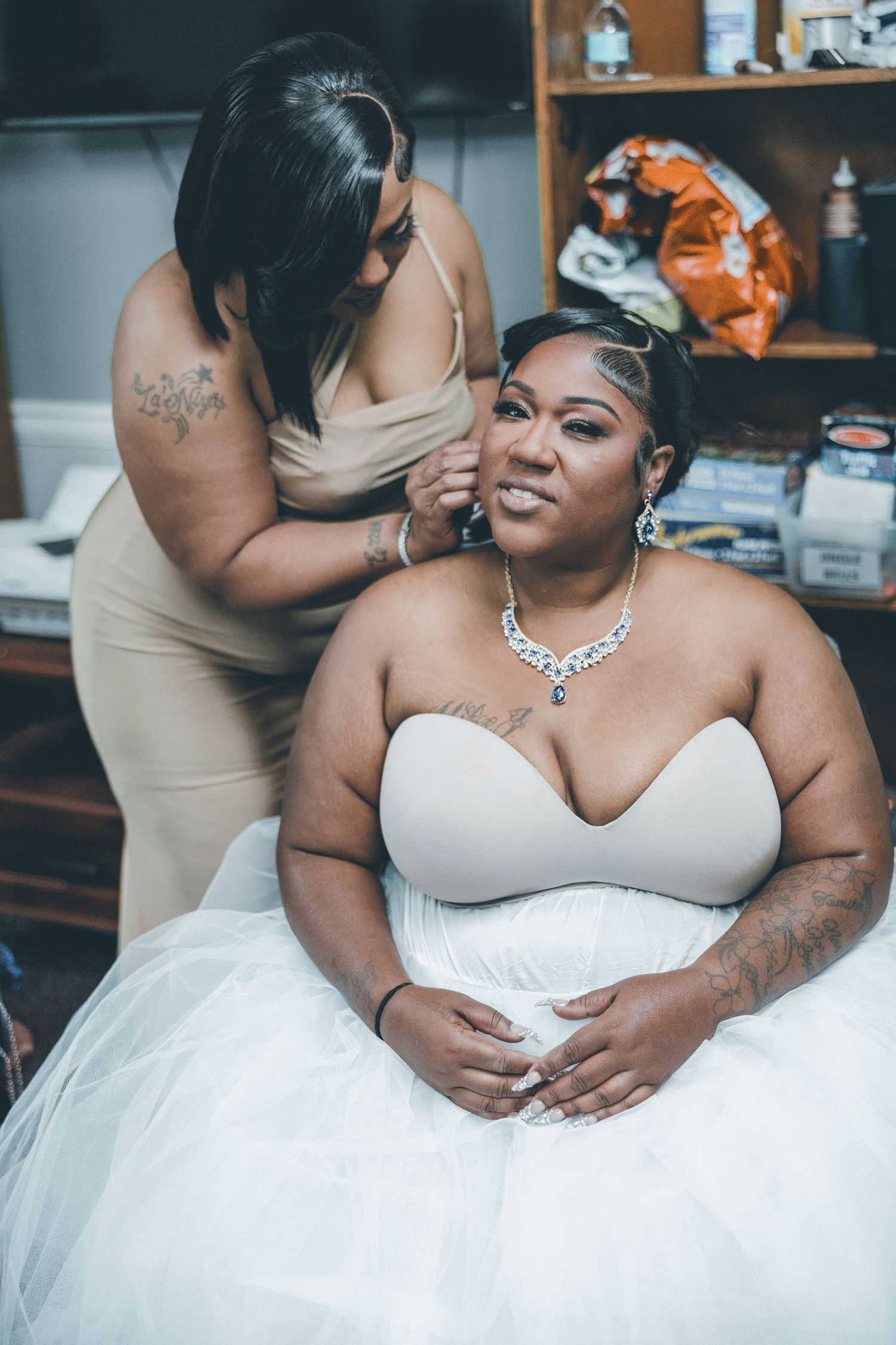 Bride in strapless wedding gown and diamond necklace gets earring help from bridesmaid during wedding preparations.