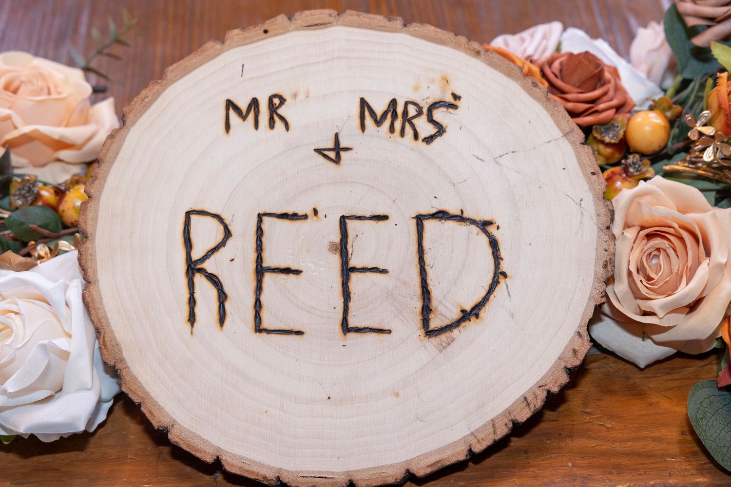 A rustic wood slice wedding sign with 'MR & MRS REED' surrounded by blush roses and greenery on a wooden surface.