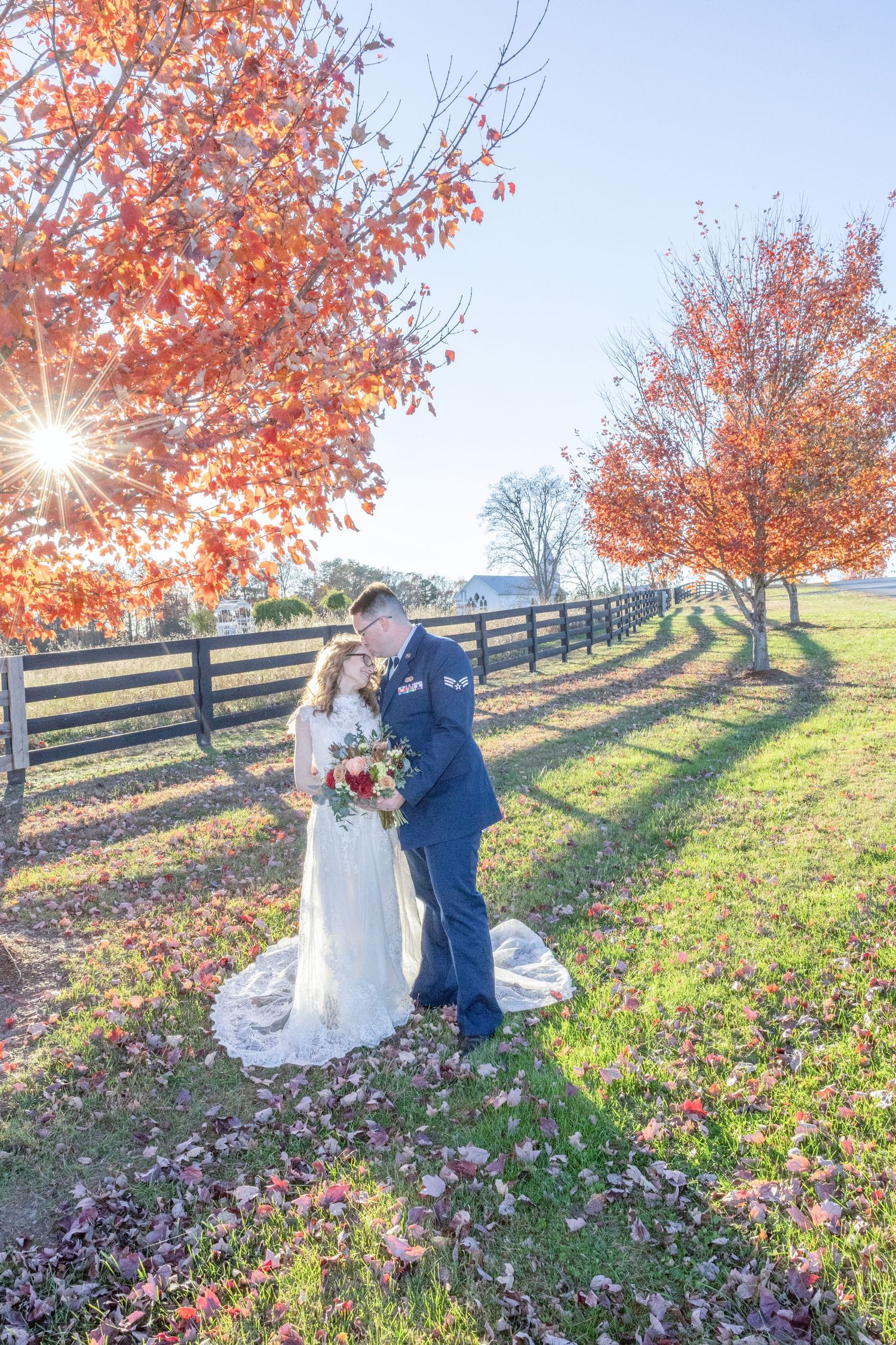 A couple embraces on a grassy hillside with autumn trees and a wooden fence during a sunny fall afternoon.