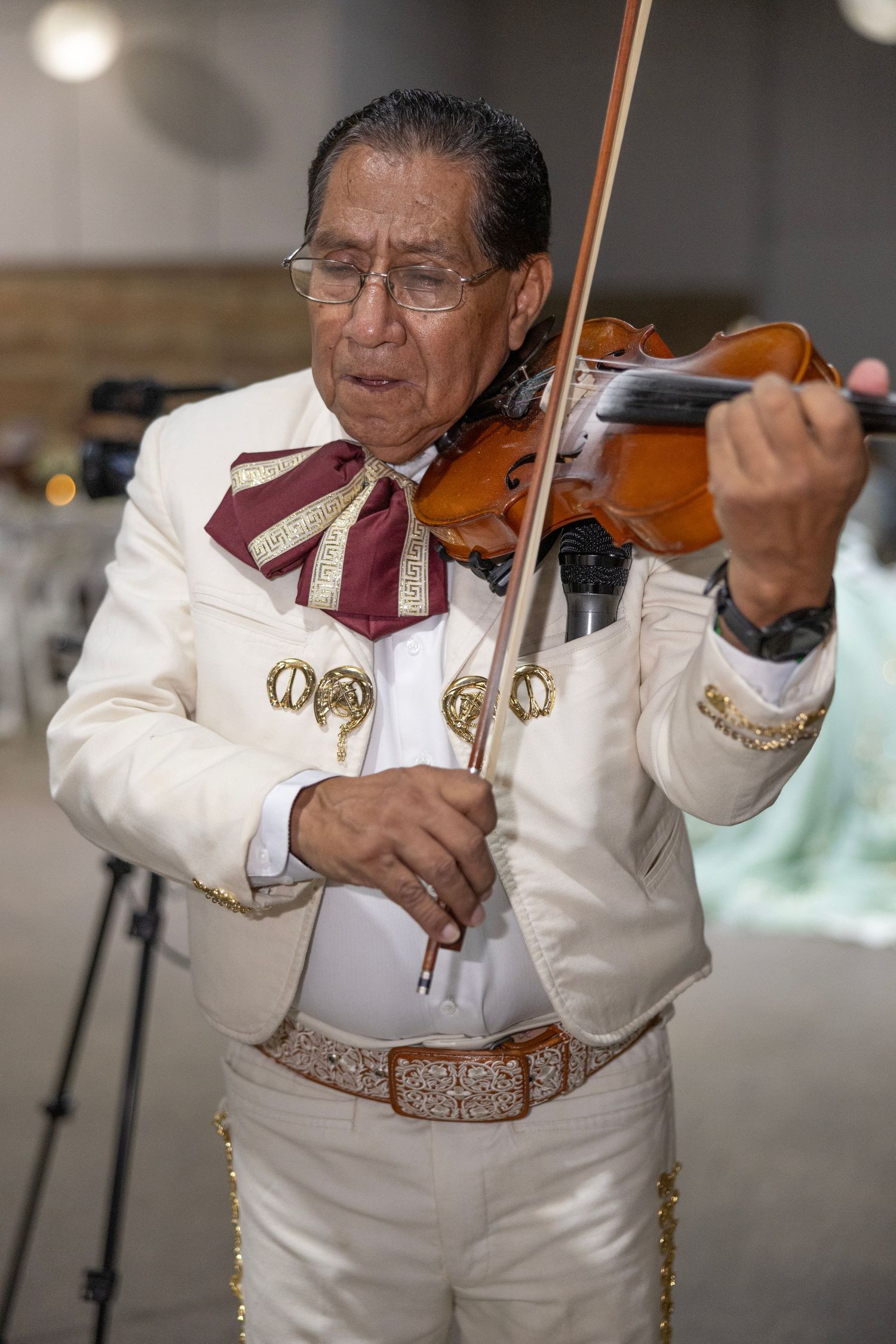 A mariachi musician in traditional white embroidered attire plays violin with focused concentration.