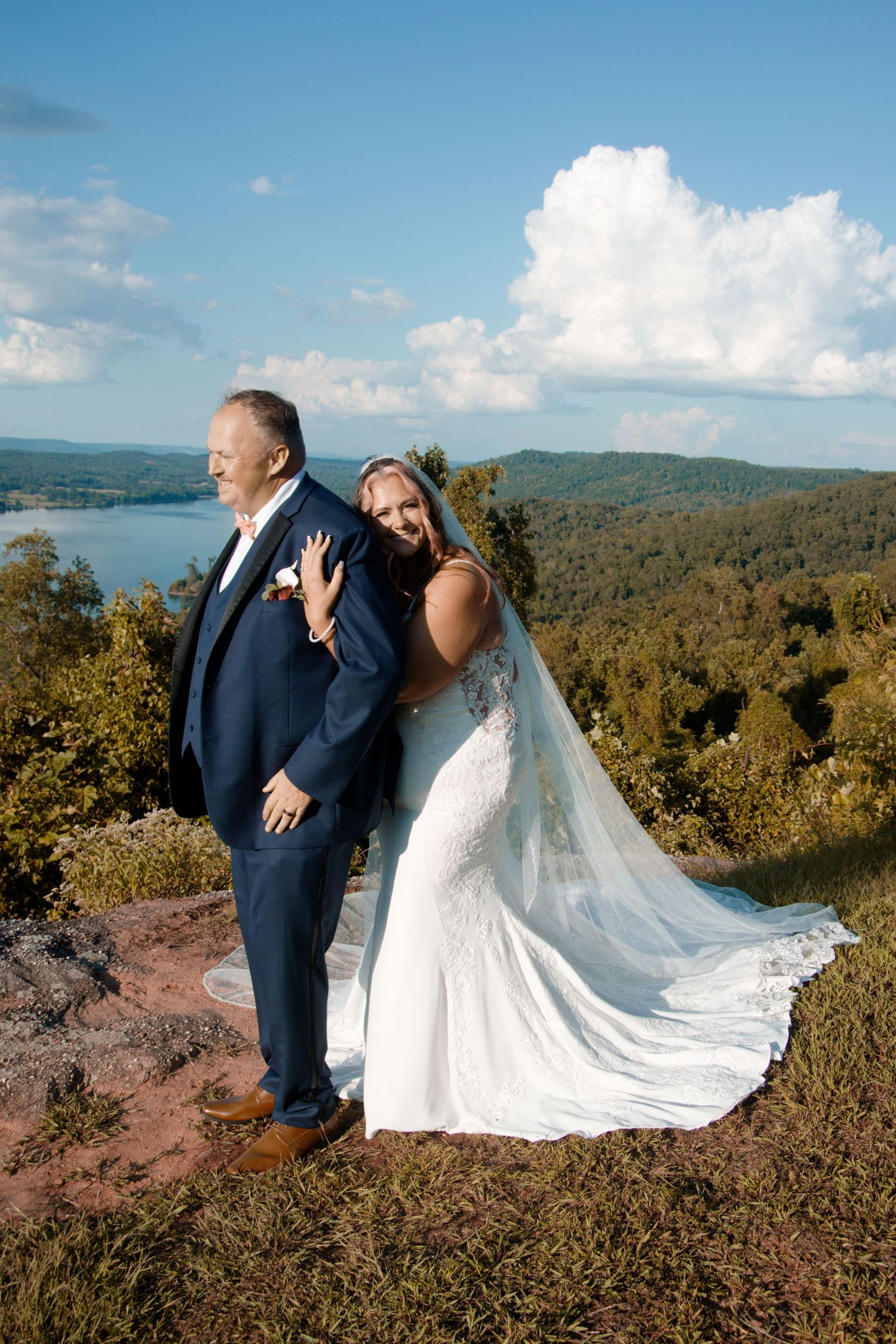 A bride and groom share romantic poses on a scenic mountain overlook with sweeping forest and lake views in the background.