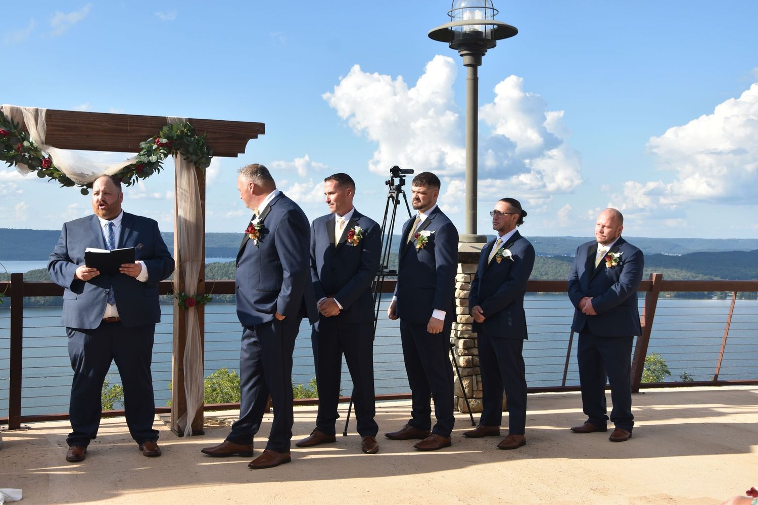 Wedding party stands in formal attire at scenic waterfront venue with panoramic ocean views and wooden pergola.