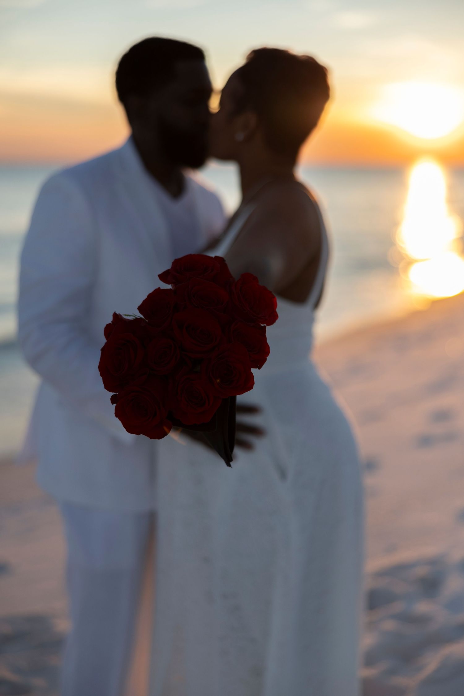 A romantic beach photoshoot silhouette of a couple in white attire holding red roses at sunset over the ocean.