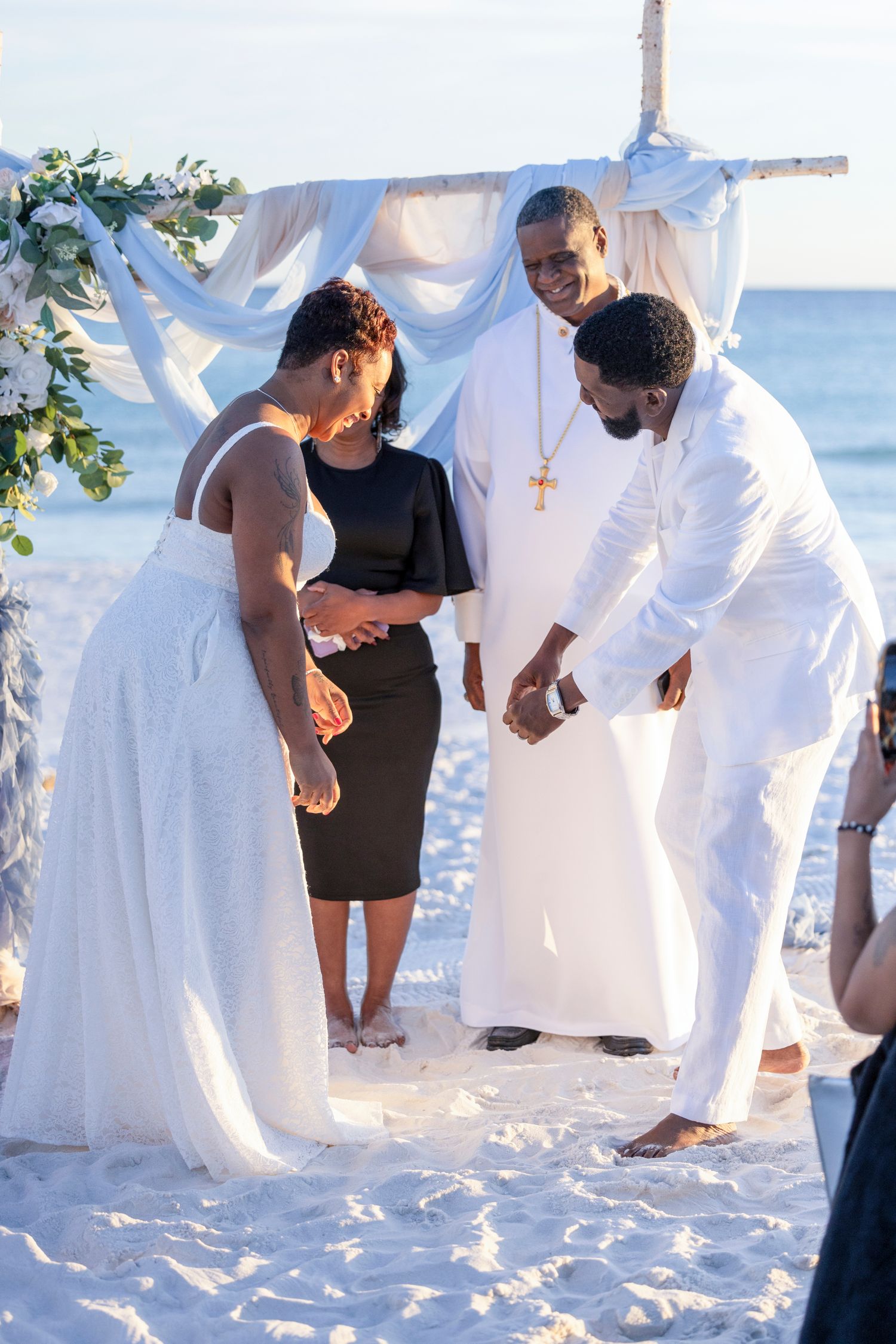 A beachside wedding ceremony takes place under a white fabric arch with floral decorations as the sun sets over the ocean.
