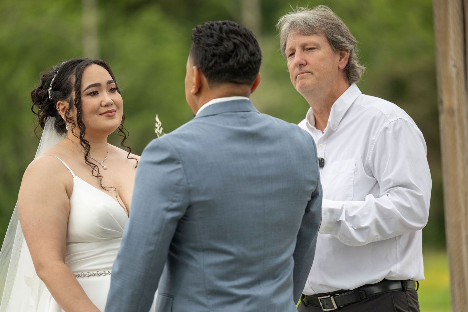 A couple exchanges vows during an outdoor wedding ceremony with an officiant in a white shirt.