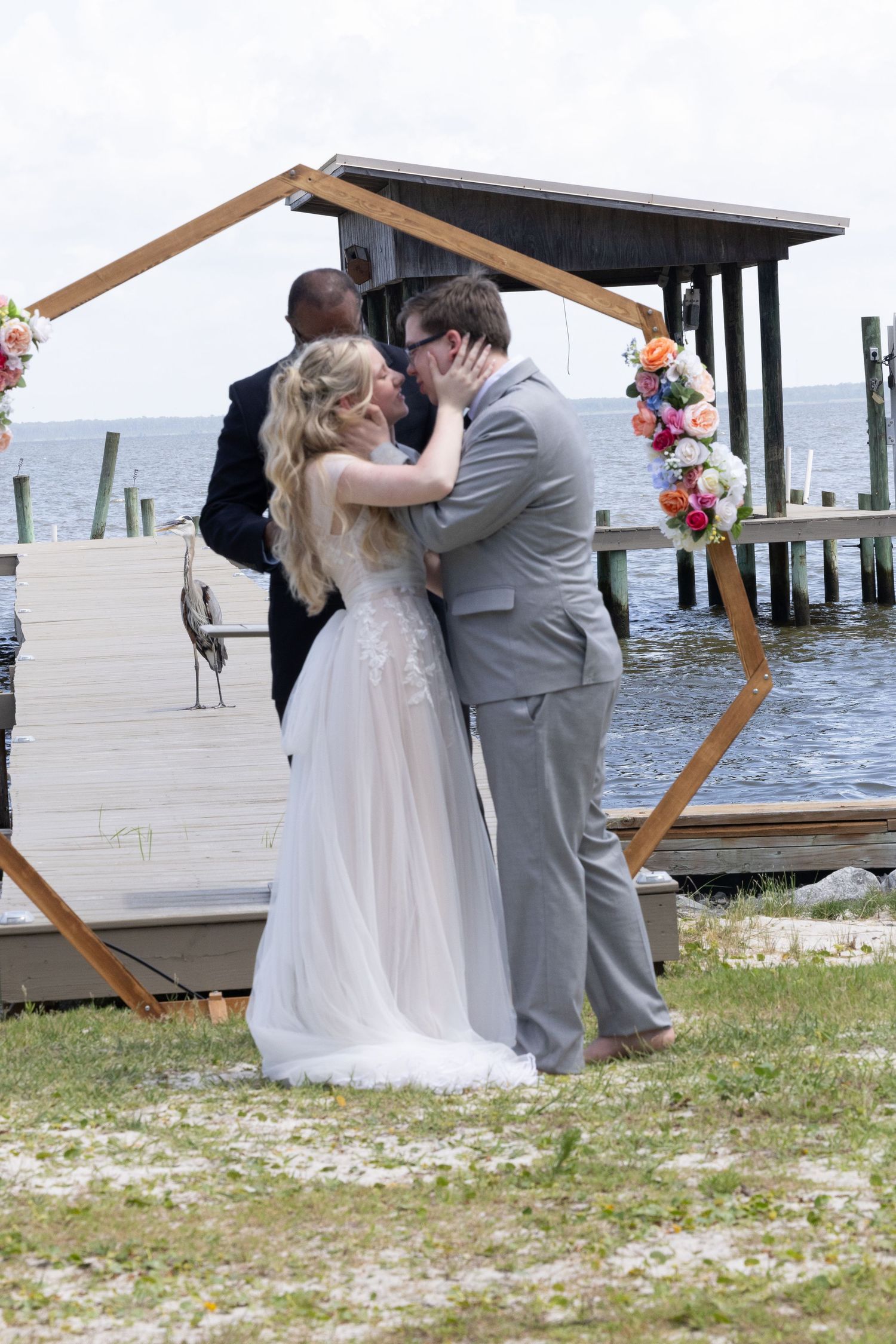 A couple shares their first kiss during a waterfront wedding ceremony under a wooden hexagonal arch decorated with flowers.
