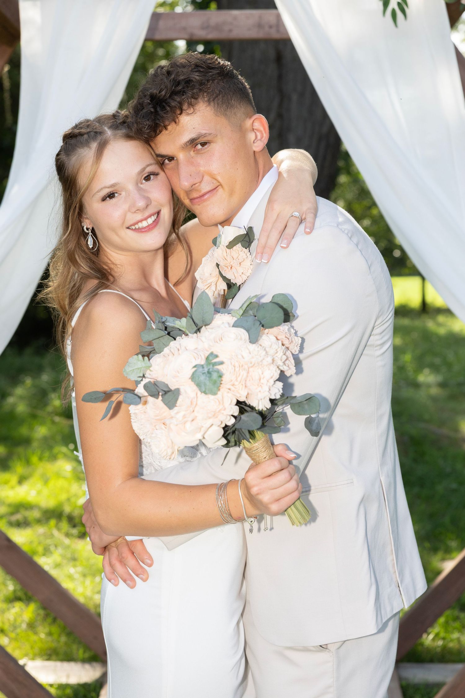 A couple in white wedding attire share intimate moments under a draped outdoor canopy with greenery in the background.