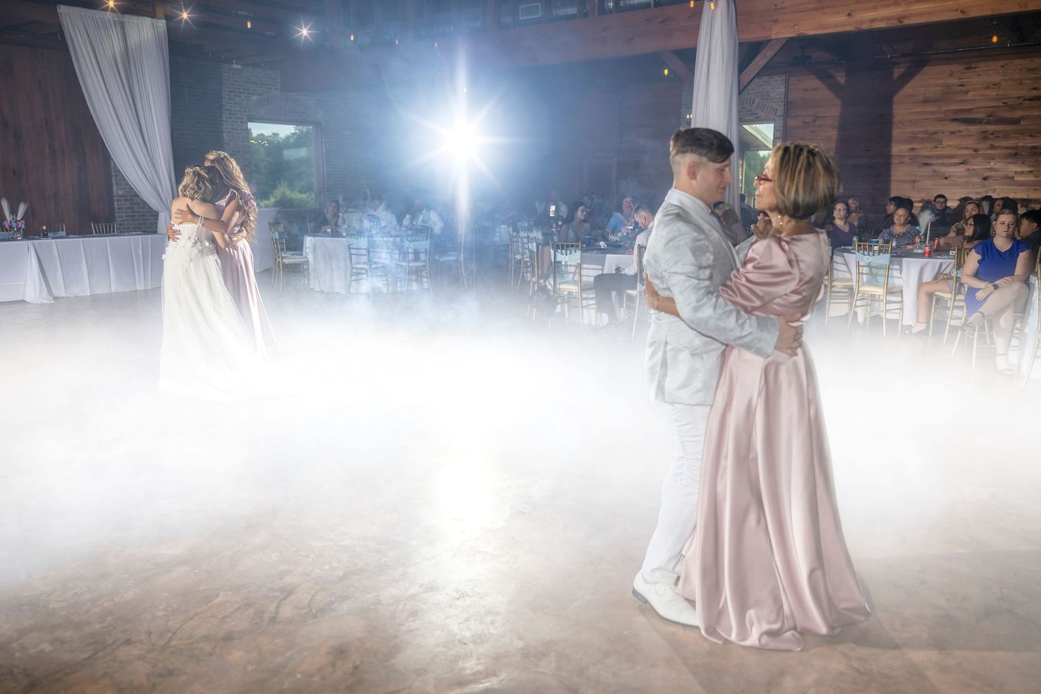 Couples slow dance on a misty dance floor at an elegant wedding reception with white decor and ambient lighting.