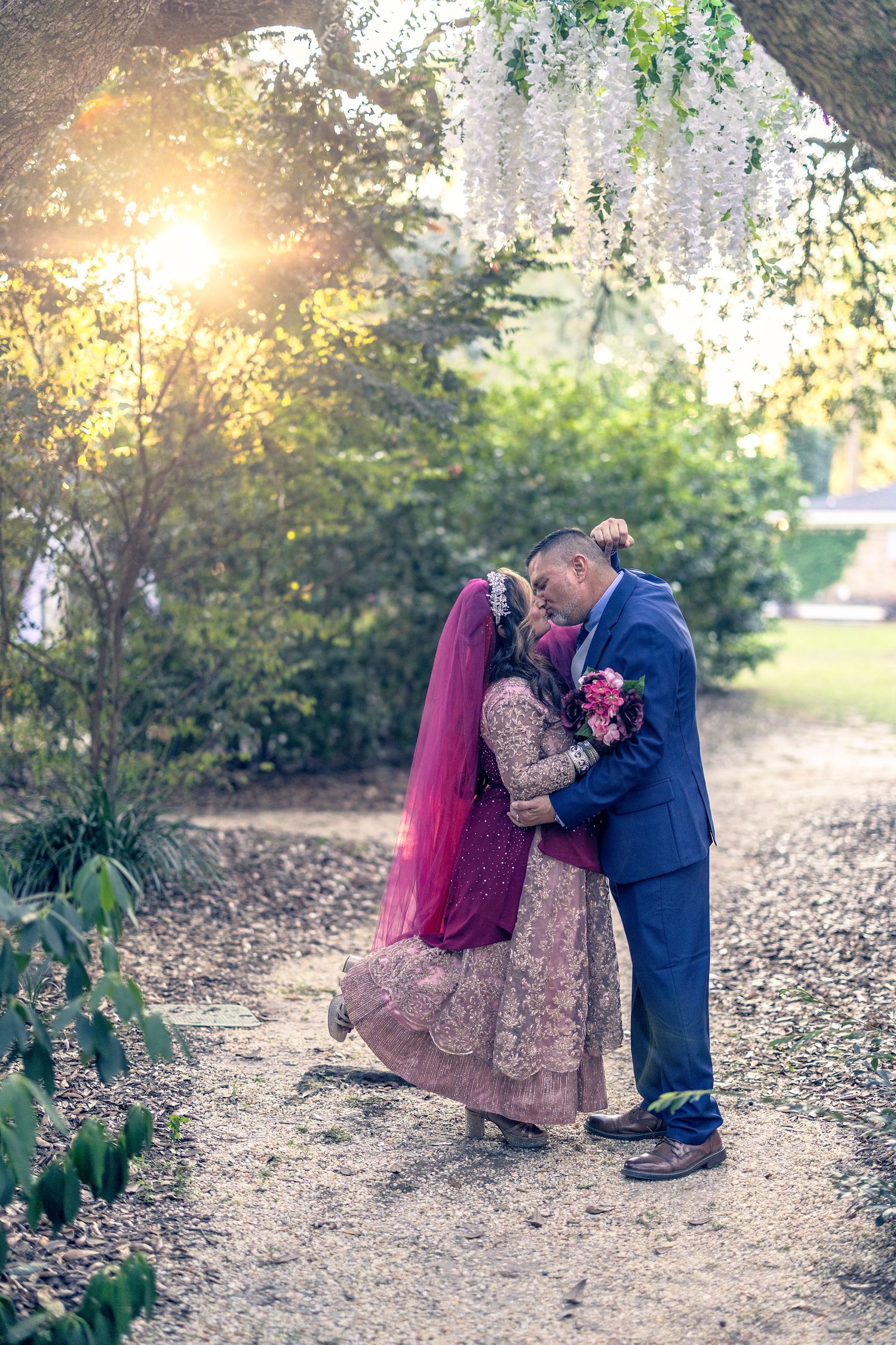 A couple shares a romantic moment under blooming wisteria at sunset in a garden path.