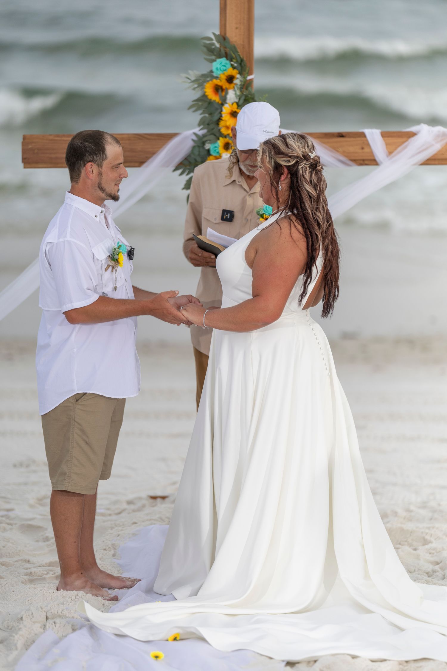 A beach wedding ceremony with a couple standing under a wooden arch decorated with yellow and blue flowers.
