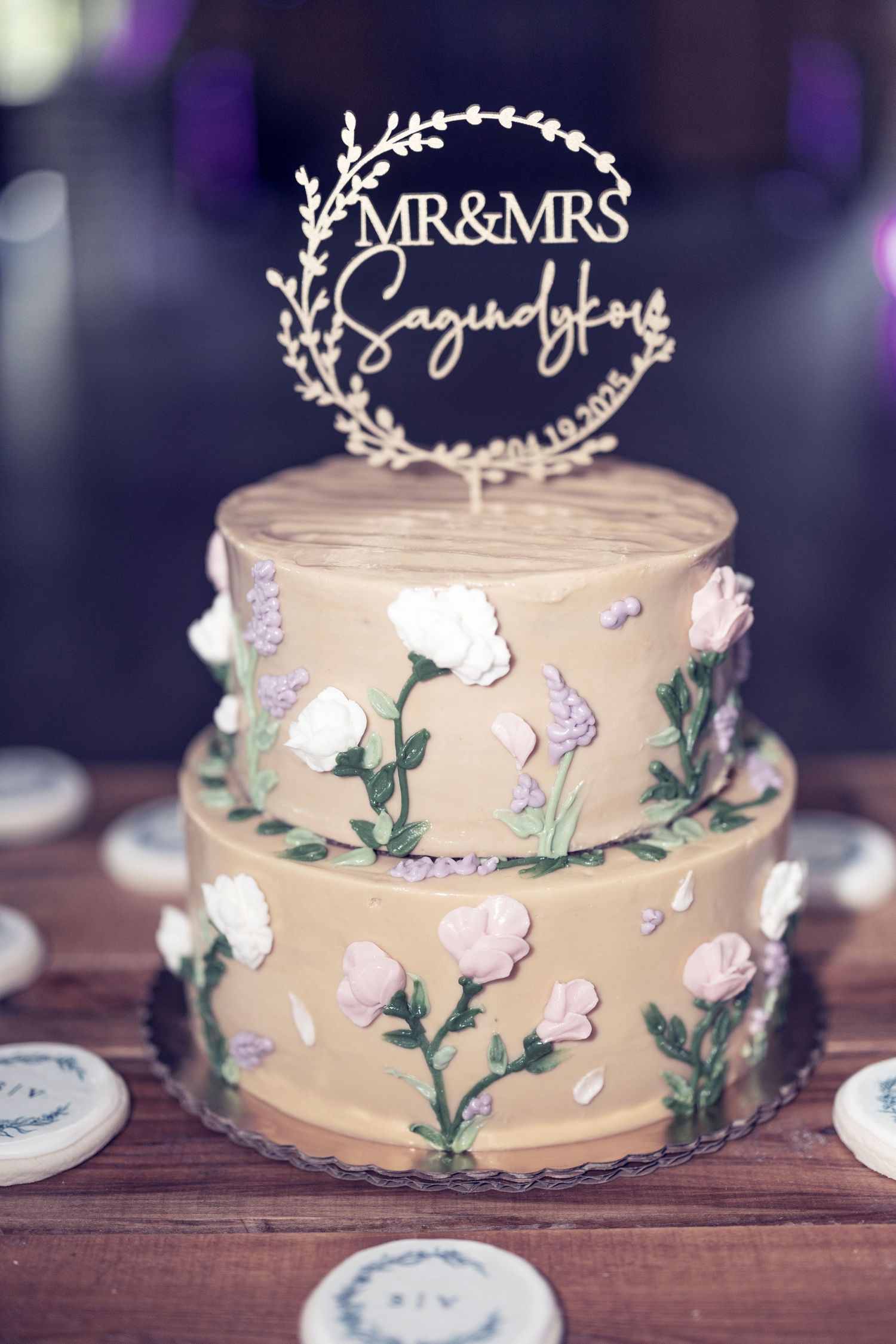Wedding cake decorated with delicate white flowers and green leaves features a 'Mr & Mrs' topper on a rustic wooden table.