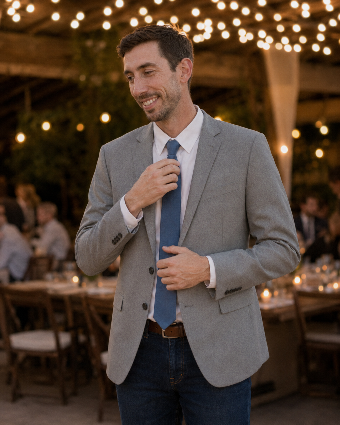 Groom in gray blazer and blue tie adjusting his tie at an outdoor wedding reception with string lights.