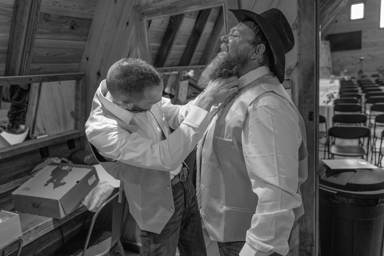 Black and white photo of two men in white shirts, one adjusting the other's tie in a rustic barn wedding setting.