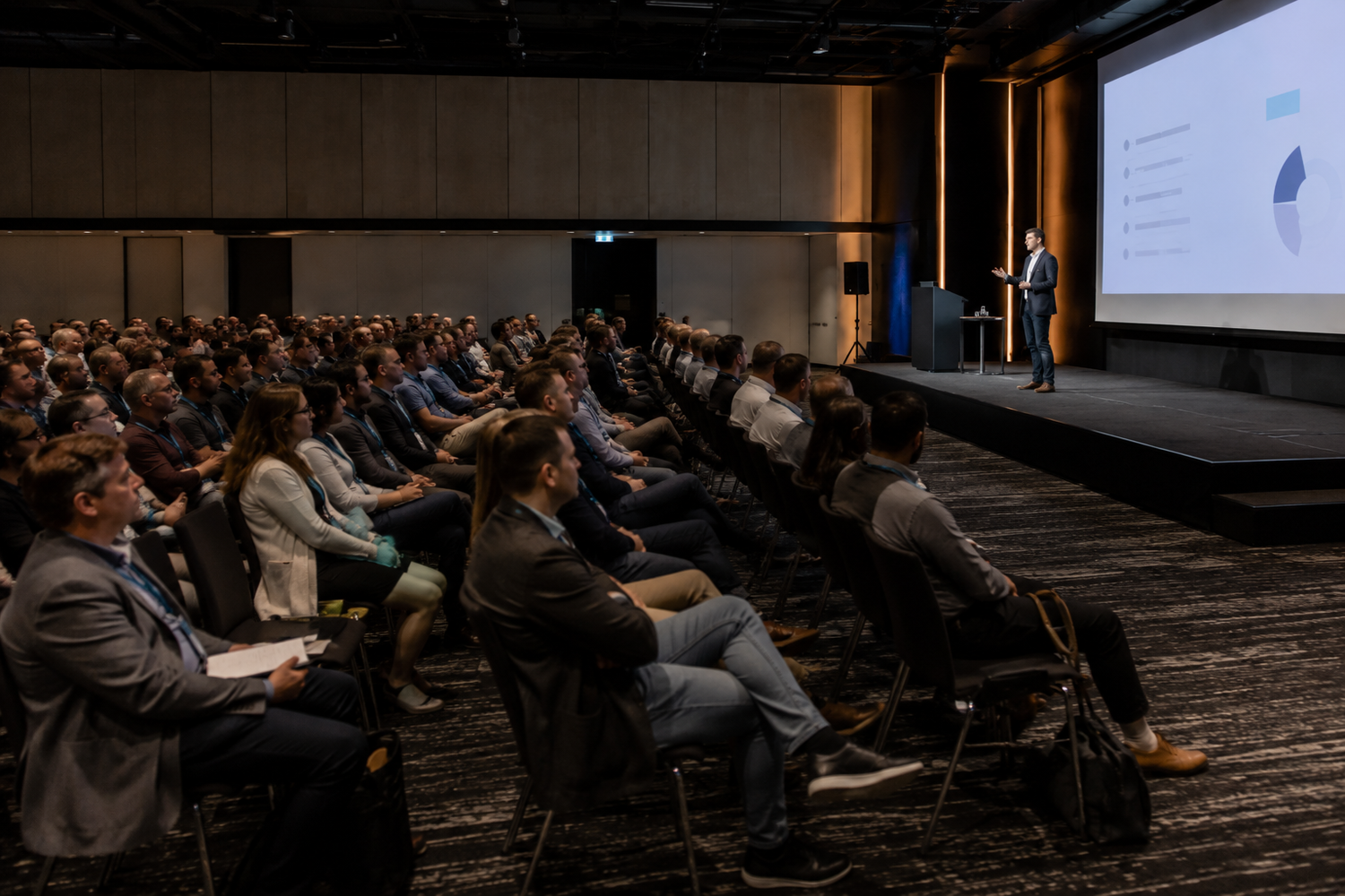 Large audience seated in modern conference hall listening to a presenter speaking at a podium on stage.