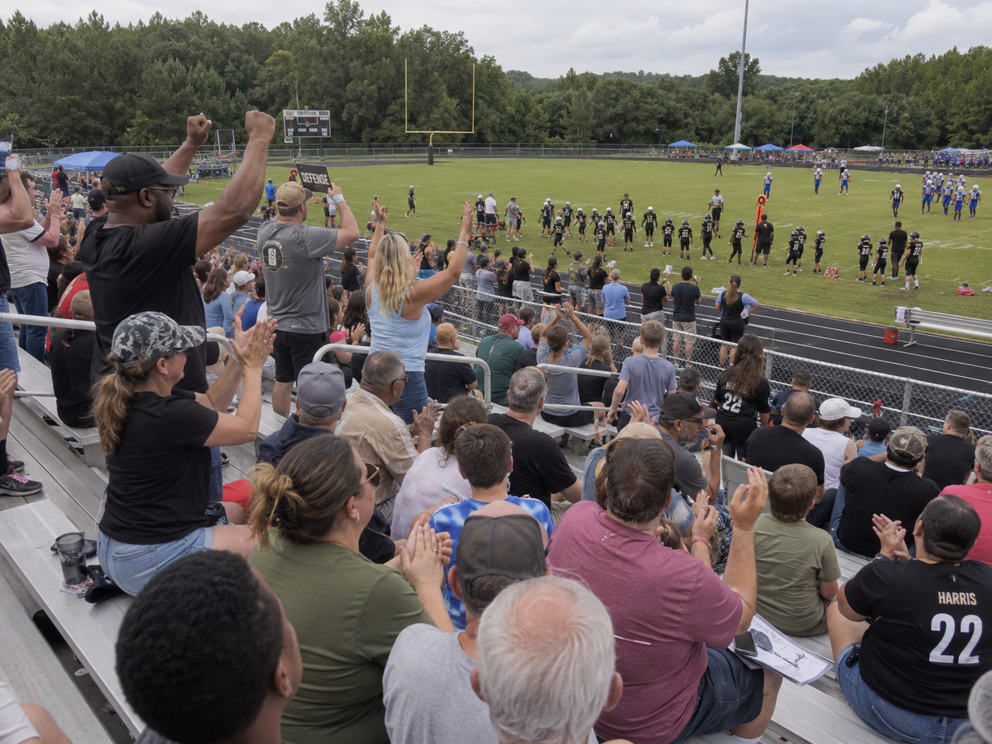 Excited crowd of fans cheering and raising fists in bleachers during an outdoor youth football game.