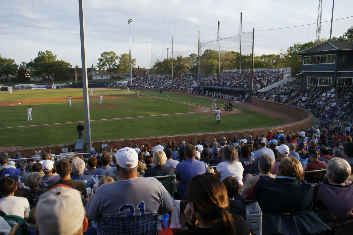 Packed crowd watches a baseball game at a sunny minor league ballpark with green trees and light towers in the background.