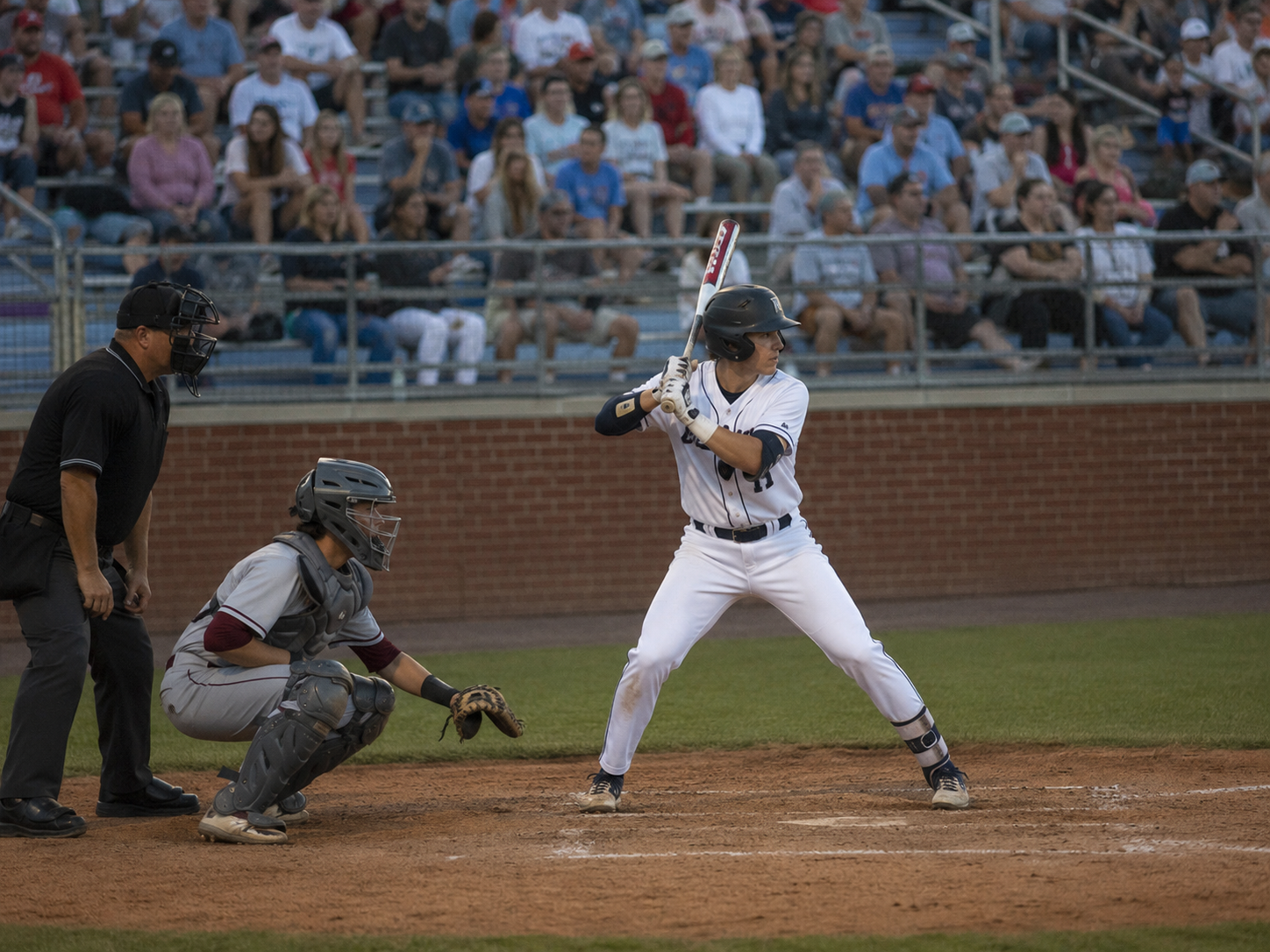 Baseball batter in white uniform stands ready at home plate with catcher and umpire during a night game.