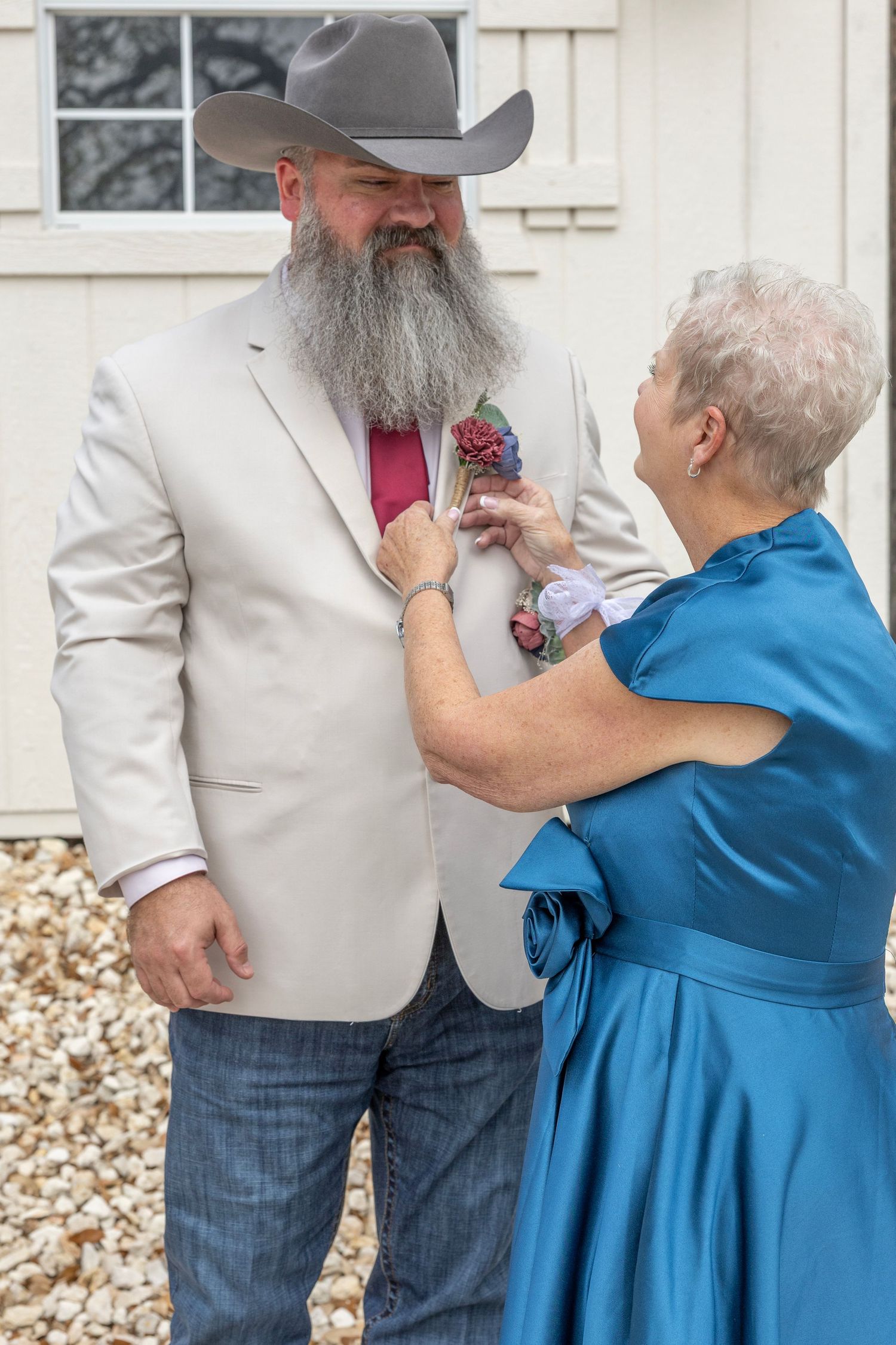 Older woman in blue dress pinning boutonniere on bearded man in cowboy hat and cream blazer at outdoor wedding.