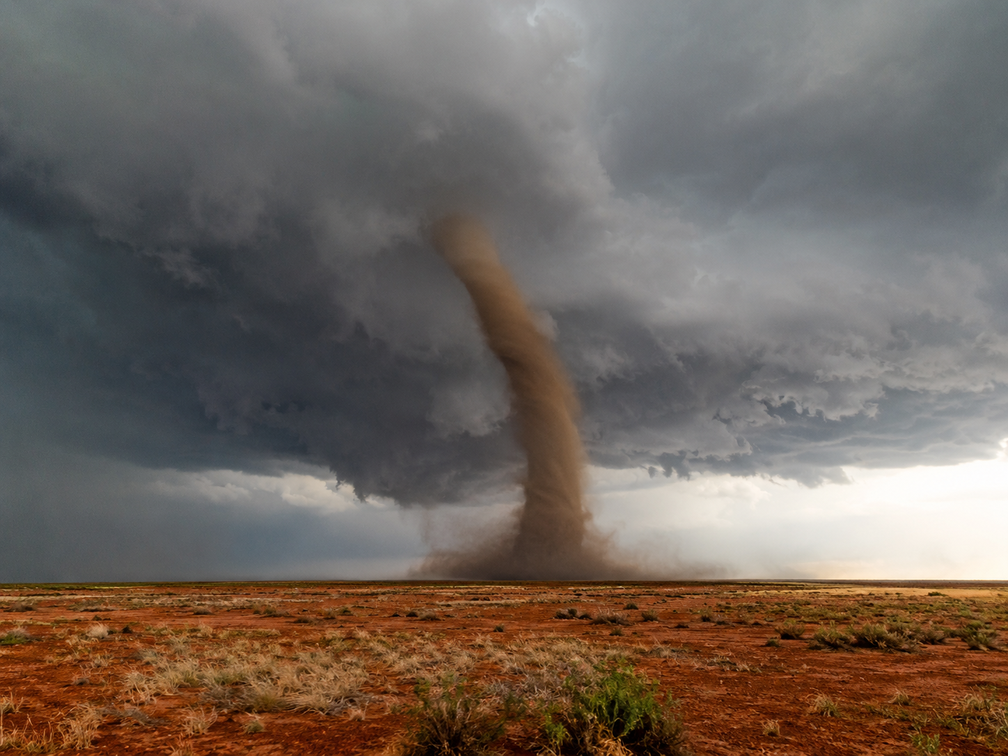 A massive tornado touches down on a flat arid plain under dark storm clouds in a dramatic natural disaster scene.