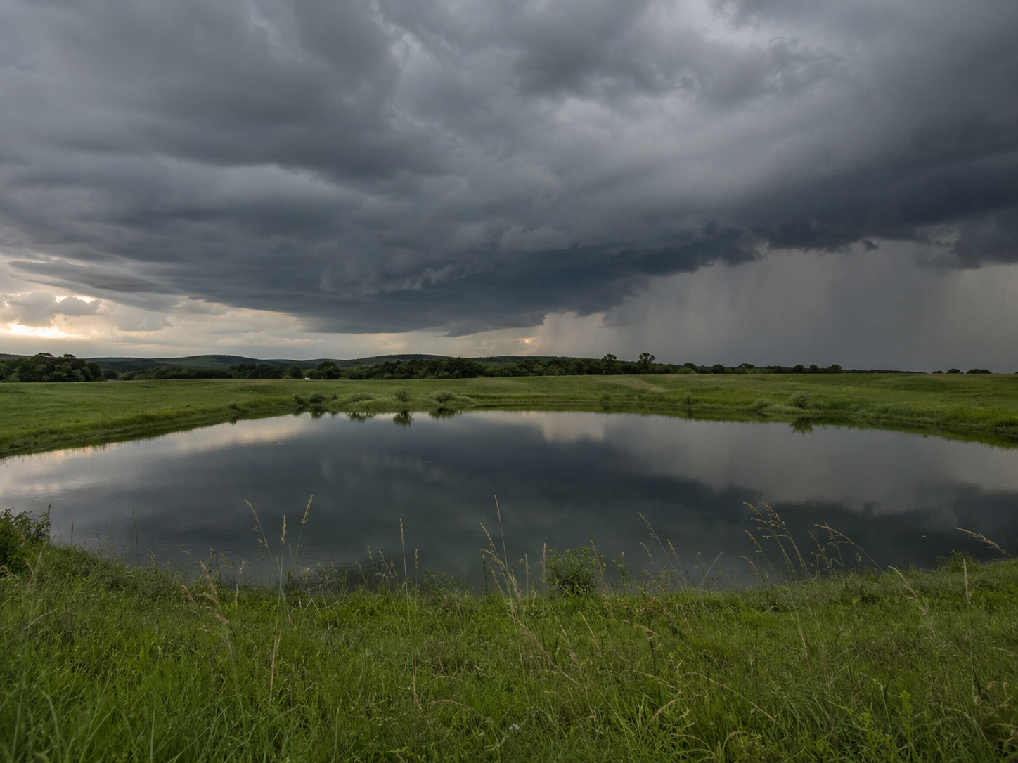 Storm clouds reflecting in a calm rural pond surrounded by lush green fields with rain visible in the distance.