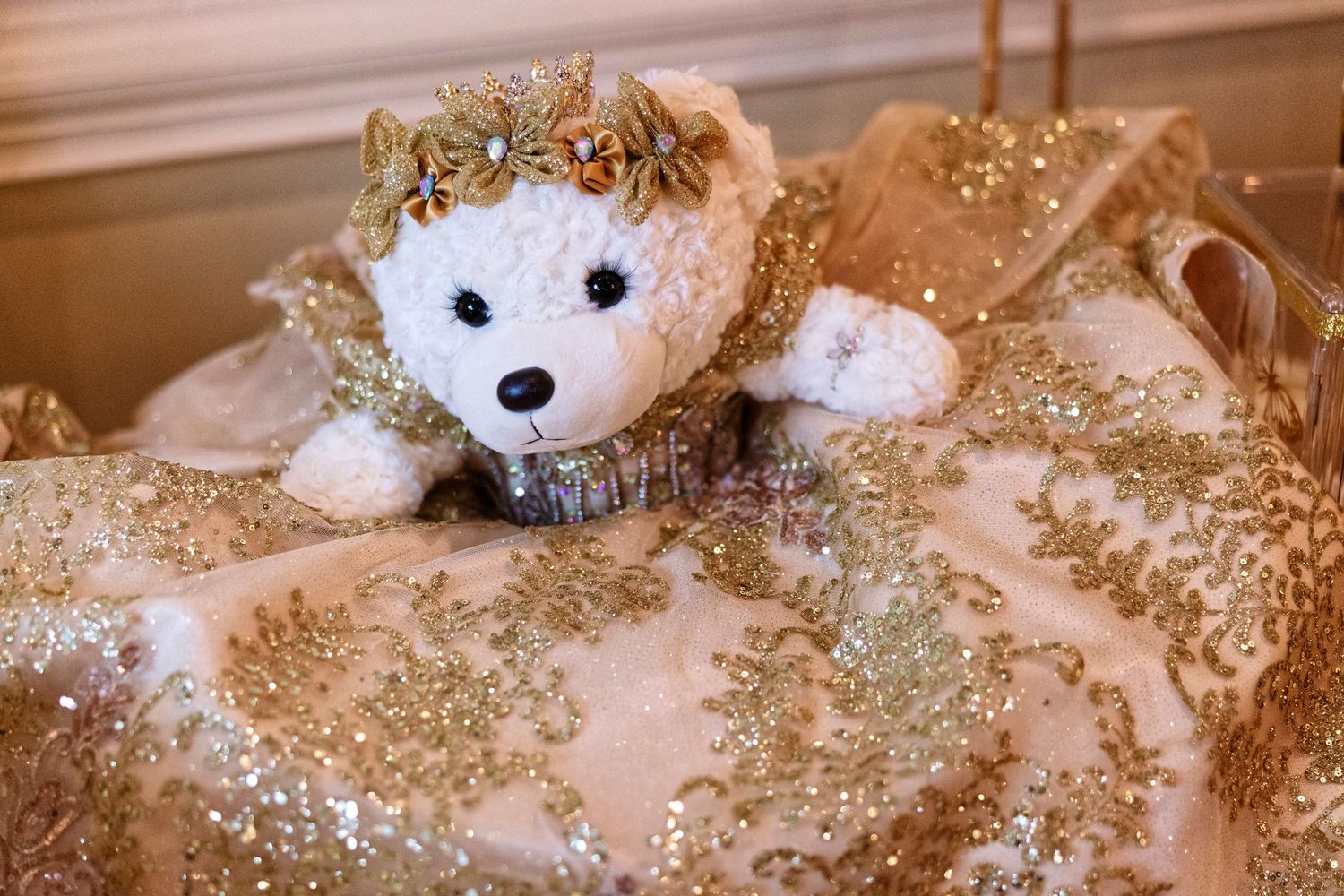 A white teddy bear wearing a gold crown sits on an elegant gold beaded and sequined fabric gown.