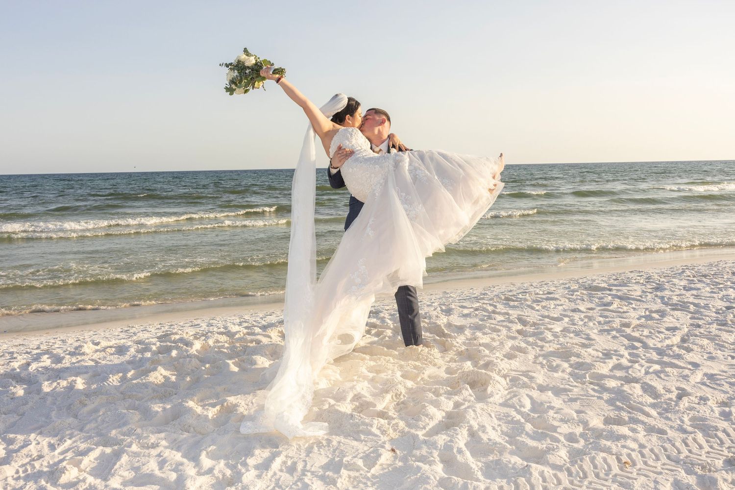 Groom lifting bride in white gown on sandy beach with ocean waves, romantic wedding photo at sunset.