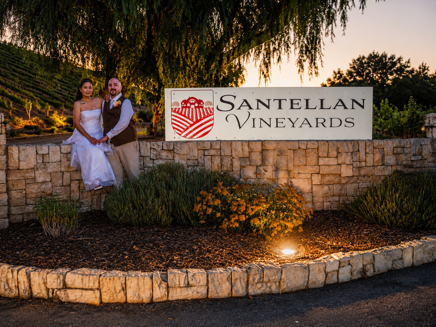 Newlywed couple posing by the Santellan Vineyards entrance sign at dusk in a romantic outdoor setting.