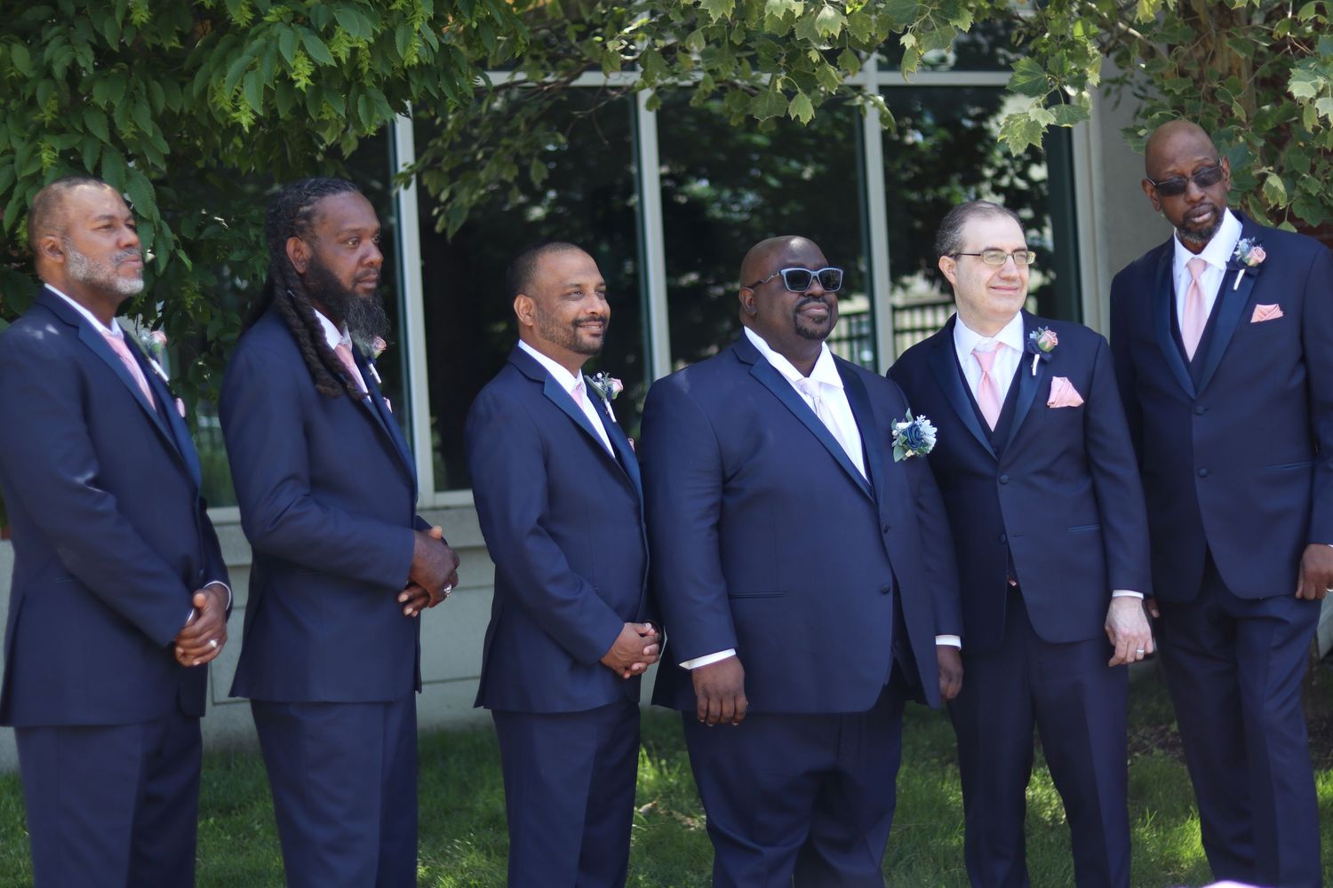 Six men in matching navy blue suits with pink boutonnieres pose together outdoors at a wedding celebration.