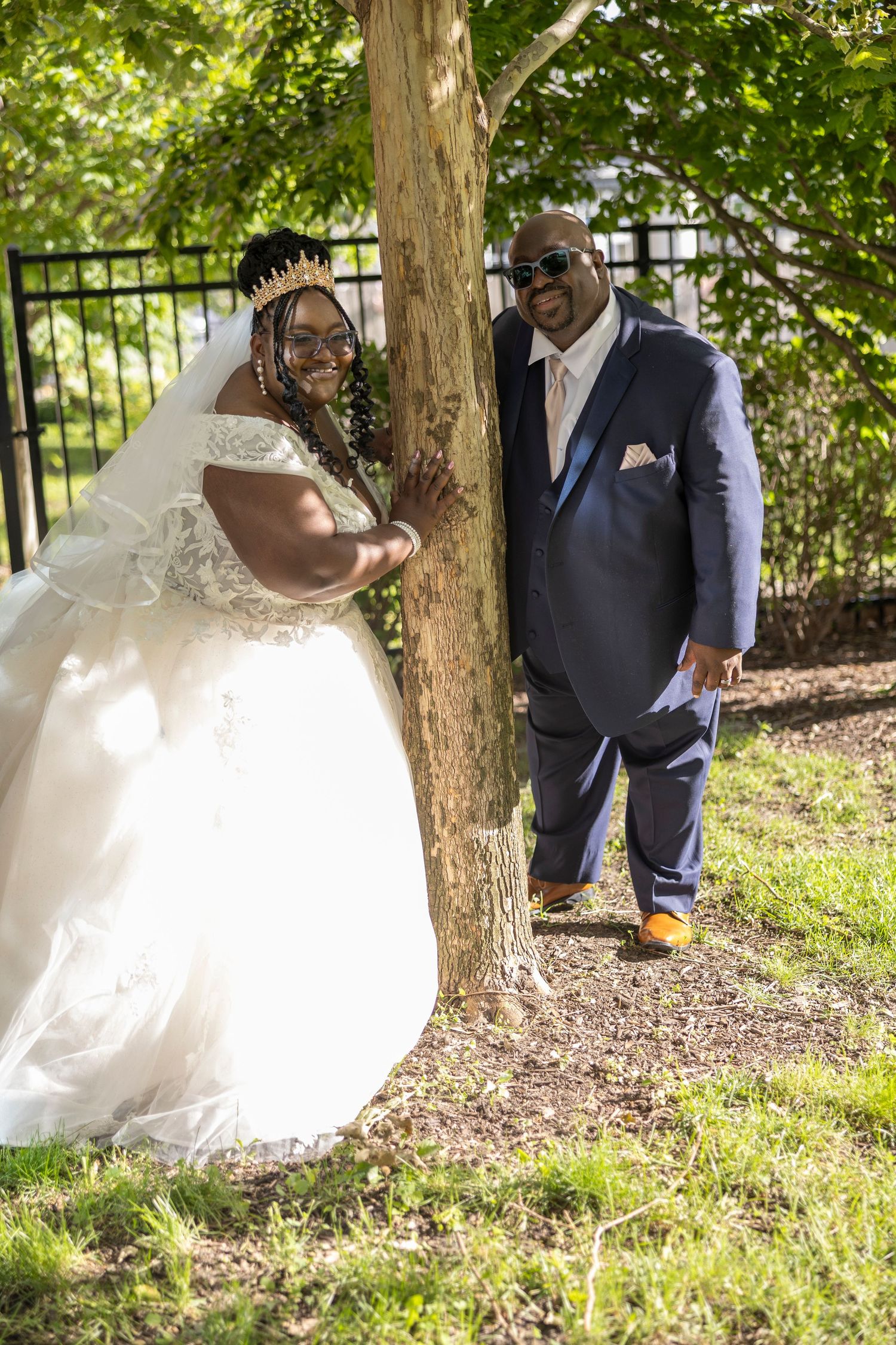 A bride in a white gown and groom in a navy suit pose by a tree outdoors on their wedding day.