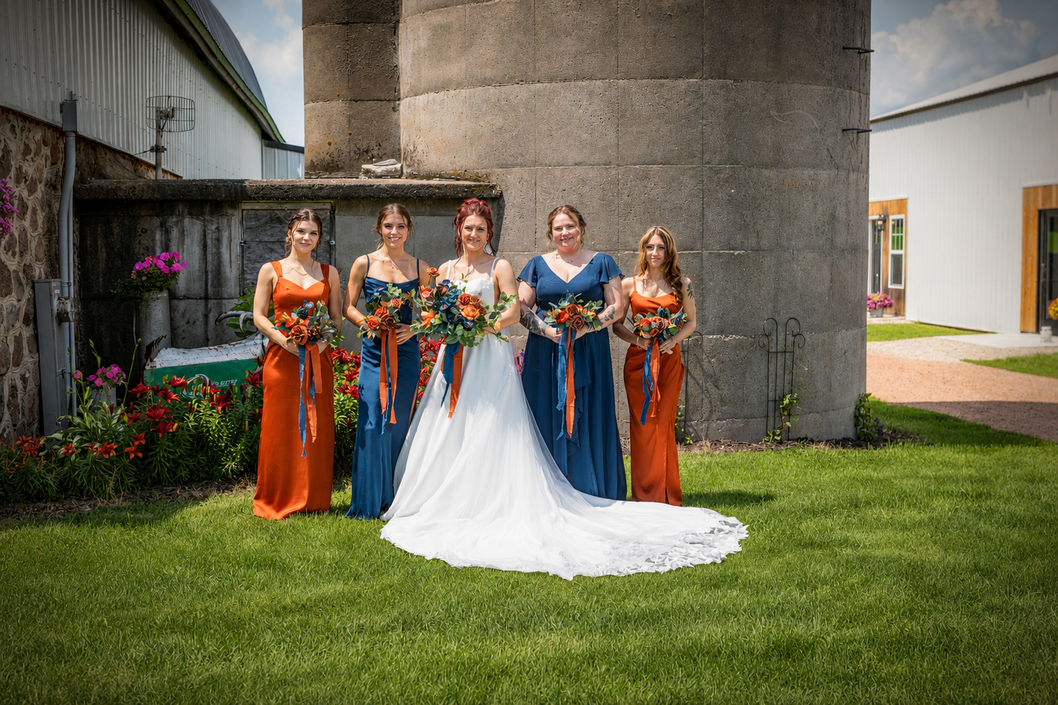 Bride with four bridesmaids in navy and orange gowns posing outdoors near a barn silo on a sunny wedding day.