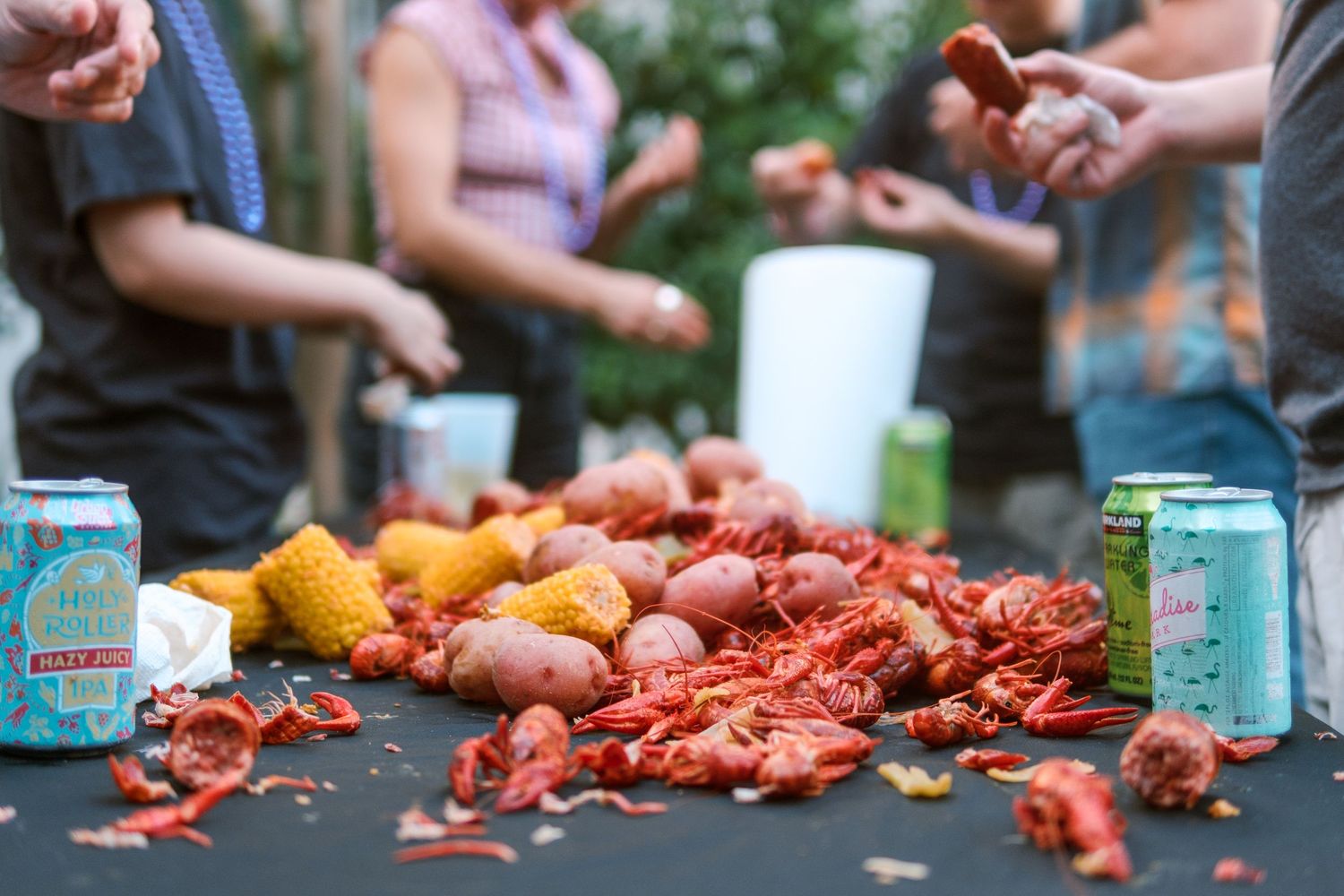 Crawfish boil spread out on table with drinks and people gathered around.