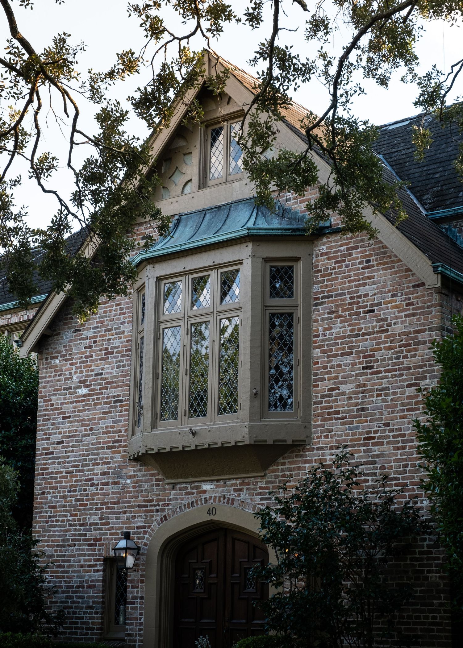 Tudor-style brick building with gothic windows and arched entryway under overhanging trees.