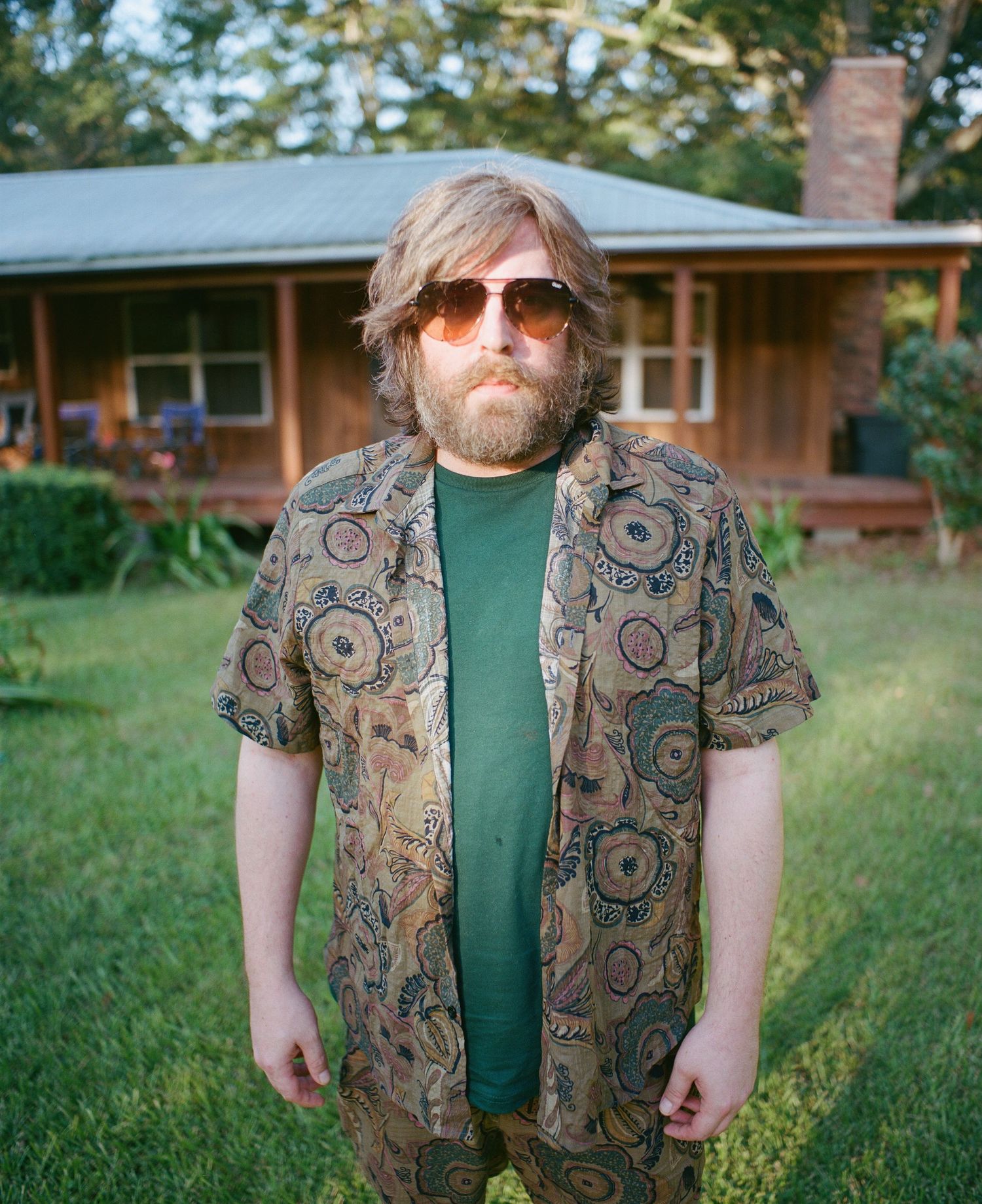 Person wearing patterned vest and green shirt standing in front of suburban house.