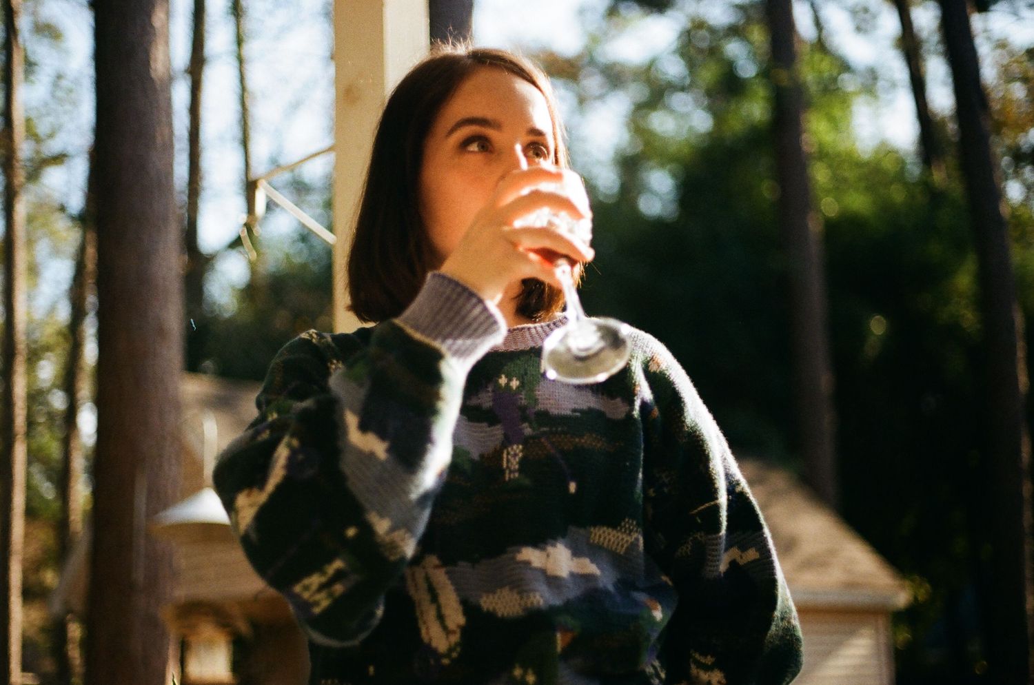 Person drinking from a glass outdoors with trees in the background on a sunny day.