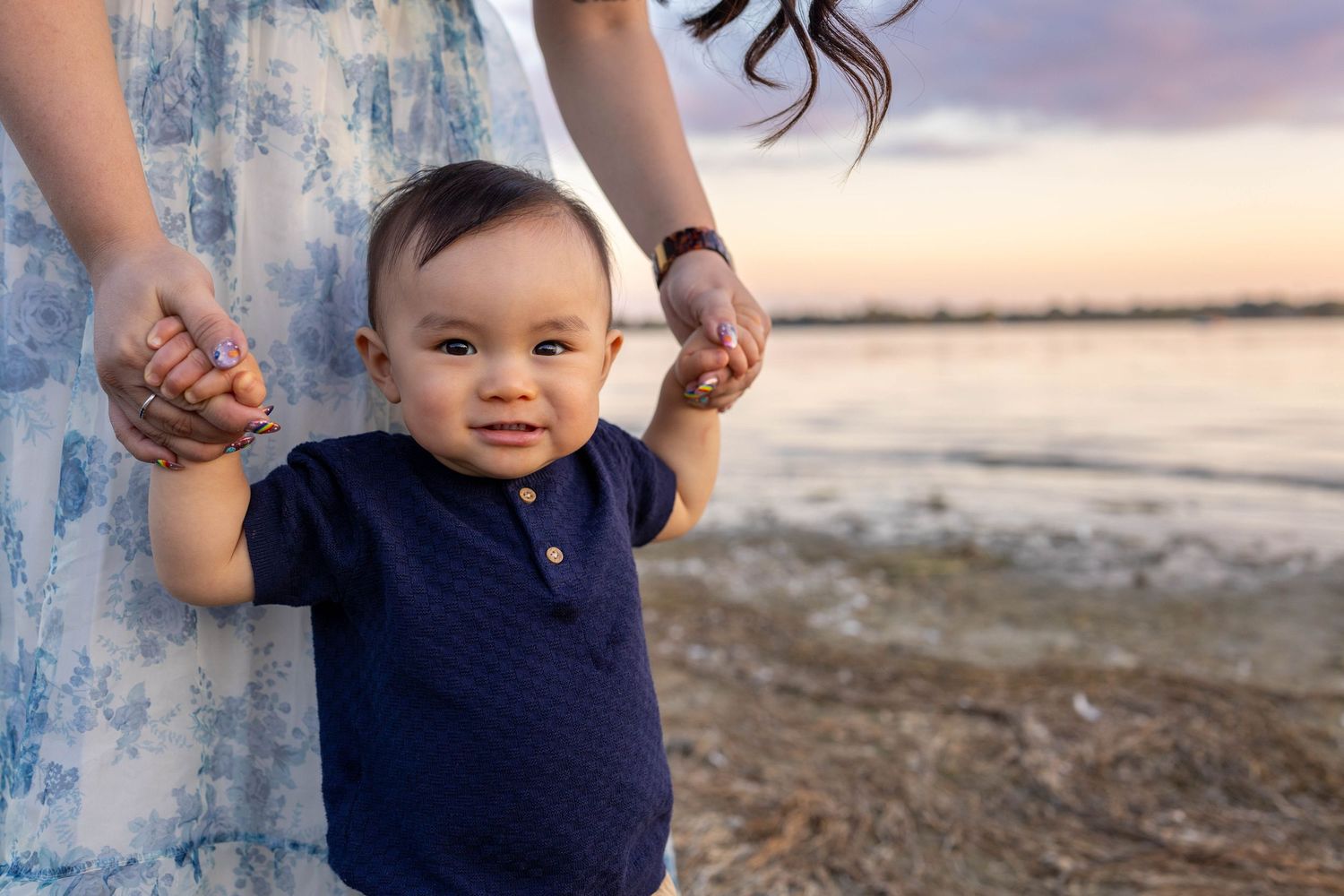 Small child smiles while holding adult hands during sunset beach photoshoot.