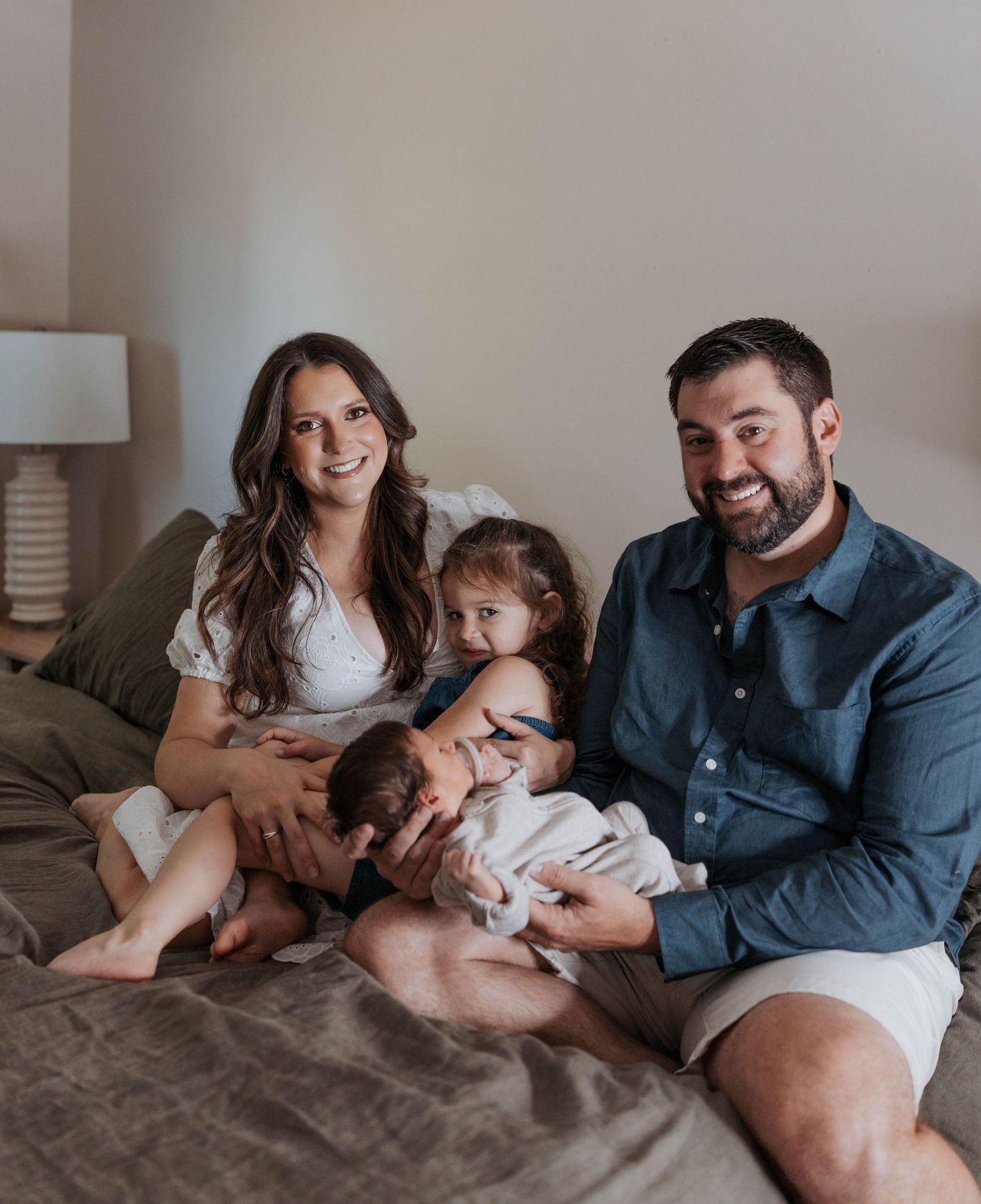 Happy family of four with mom, dad, toddler, and newborn baby posing together on a bed in a cozy bedroom.