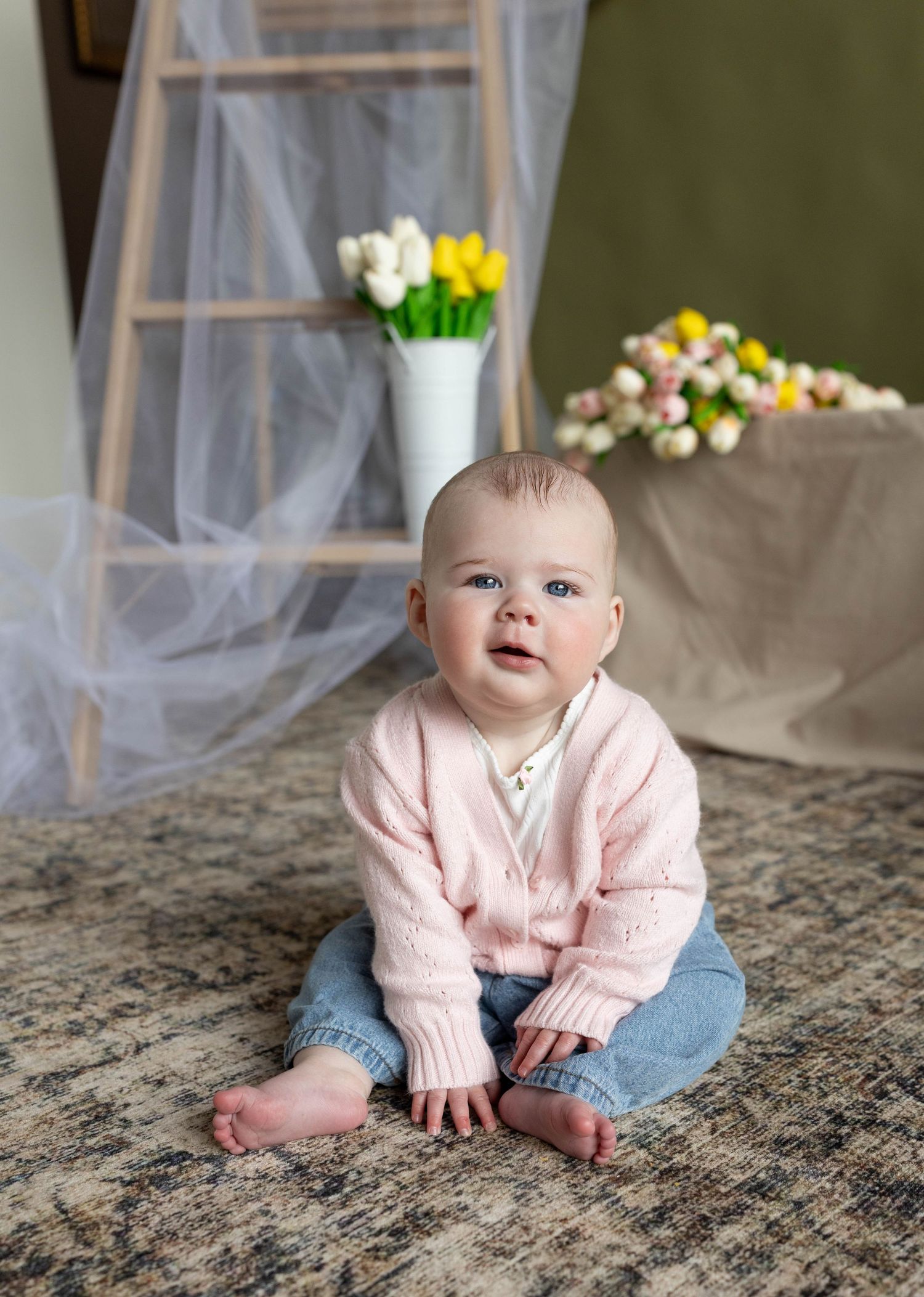 Baby girl in pink cardigan and jeans sitting on rug surrounded by spring flowers and decorative ladder.