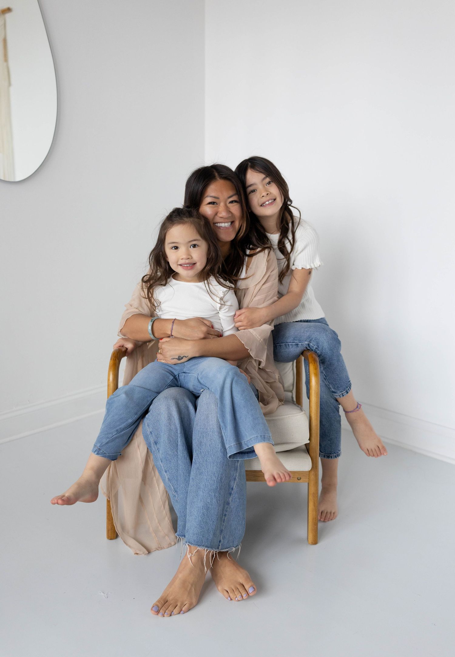 A woman and two young girls in white tops and jeans pose together on a chair in a bright white room.