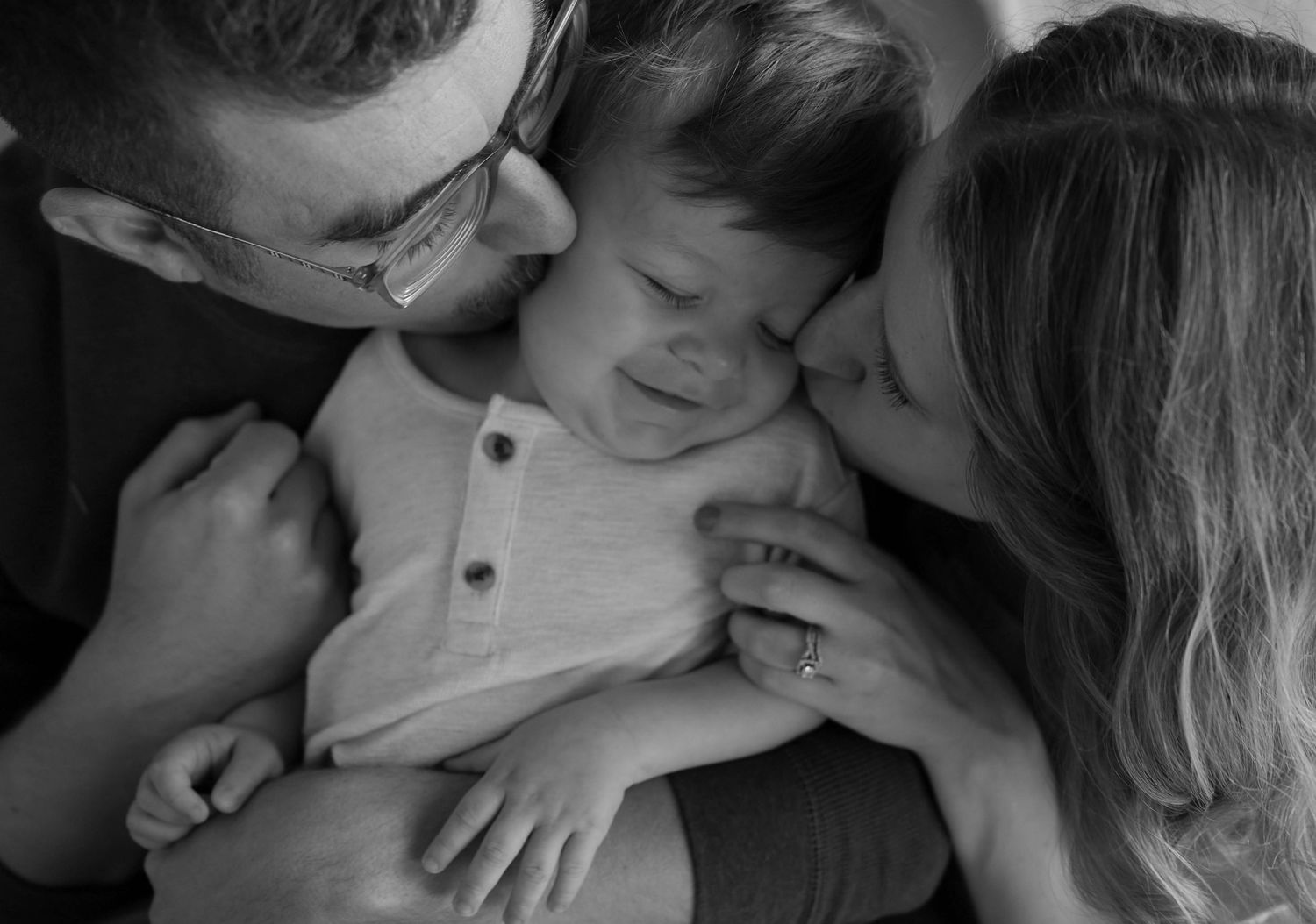 Black and white photo of parents lovingly kissing their smiling baby boy in a tender family portrait.