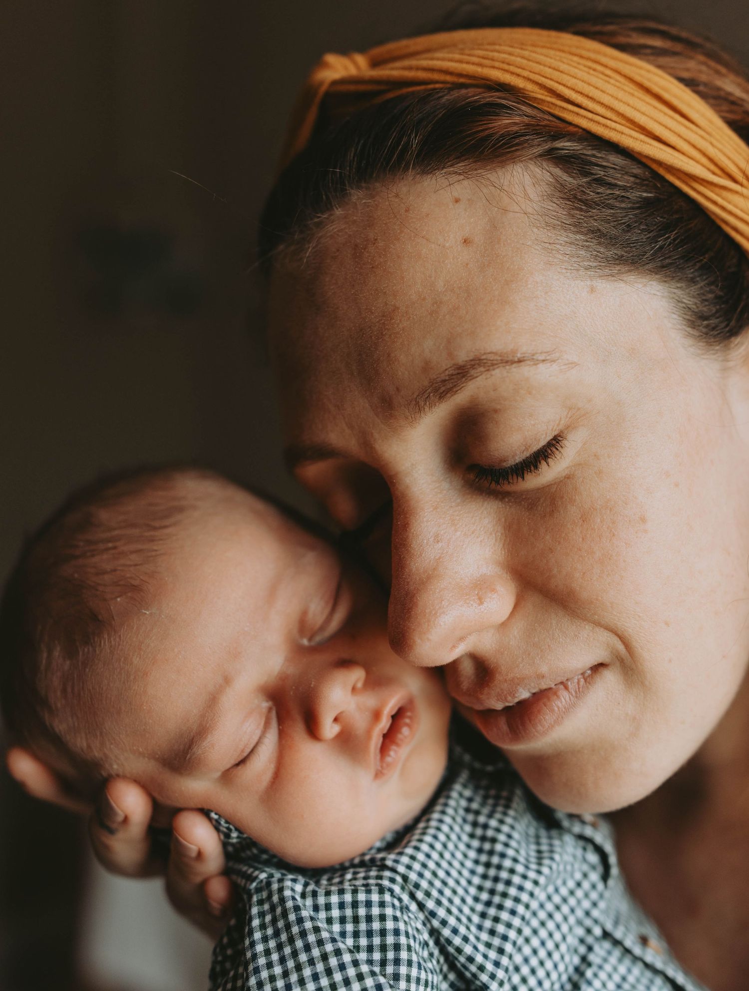 A mother with a yellow headband tenderly holds her sleeping newborn baby close to her cheek.