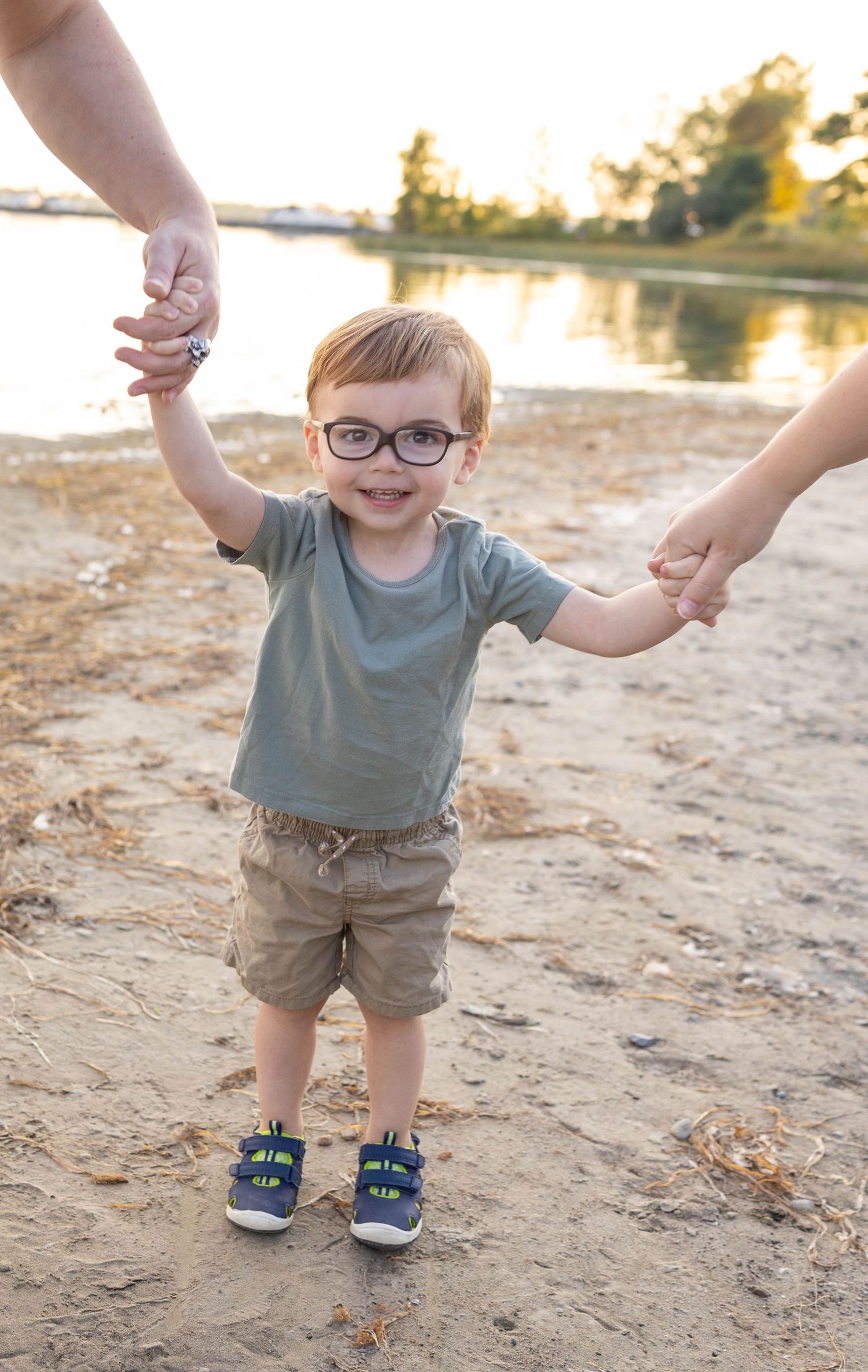 Child wearing glasses and casual clothes holds hands with adults while standing on sandy beach at sunset by lake.