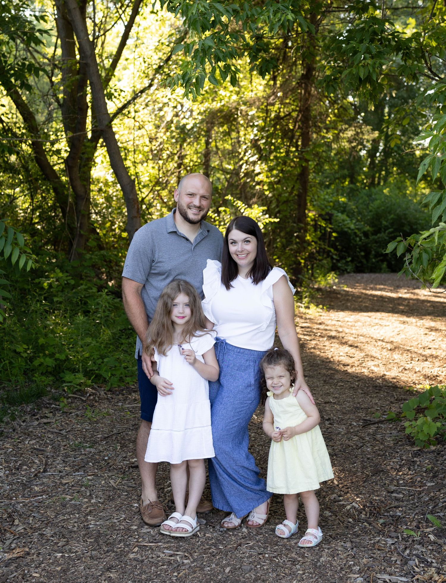 A family stands together on a sunlit forest path surrounded by lush green foliage.