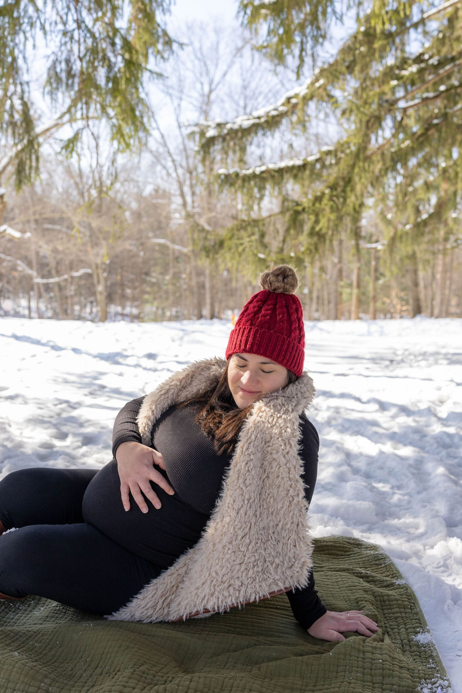 Expectant mother in red knit hat and cozy blanket sits on green blanket in snowy winter forest setting.