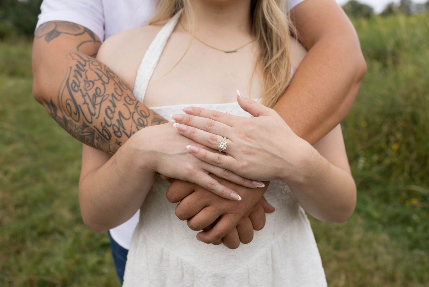 Couple embracing outdoors, woman wearing engagement ring, man with tattoed arm, in a green field setting.