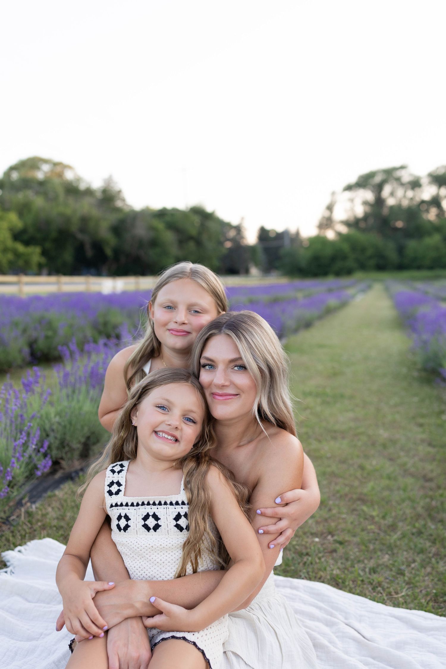 Three people in white outfits sit together on a blanket beside a beautiful blooming lavender field on a sunny day.